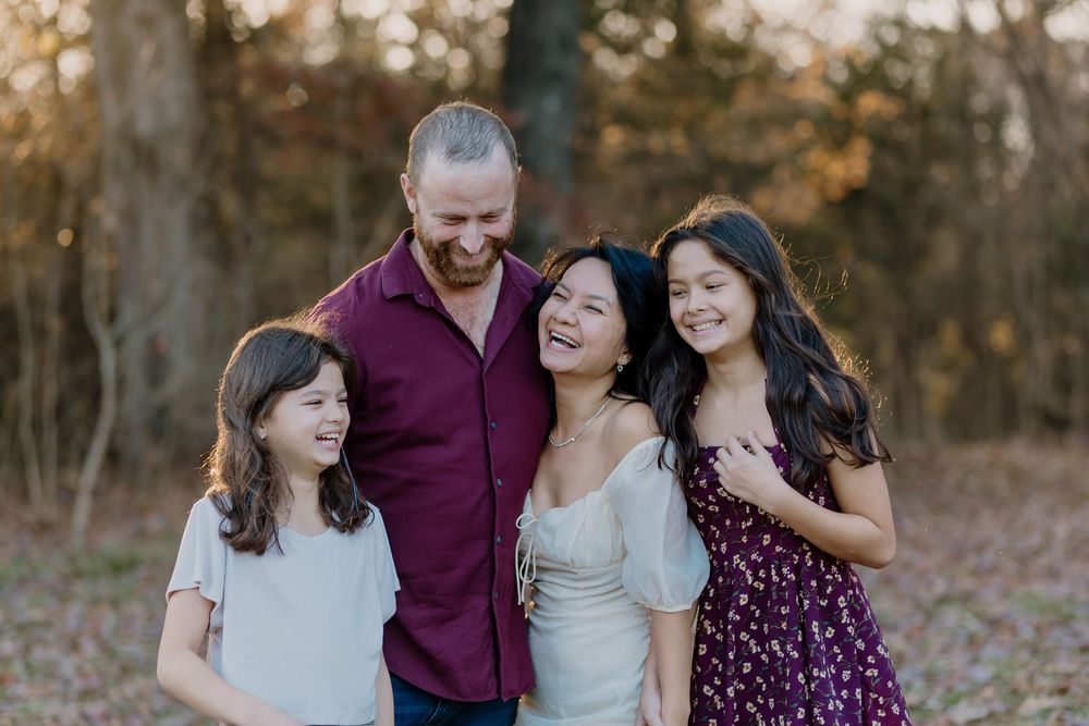 A joyful family of four laughing together outdoors in a wooded autumn setting during a professional portrait session.