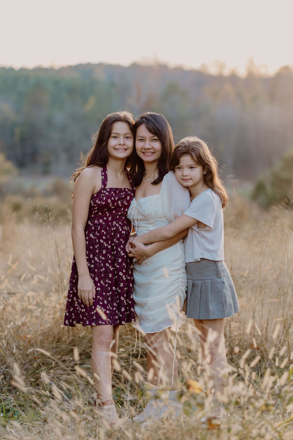 Three girls pose together in a golden field at sunset, wearing summer dresses, smiling warmly at the camera.