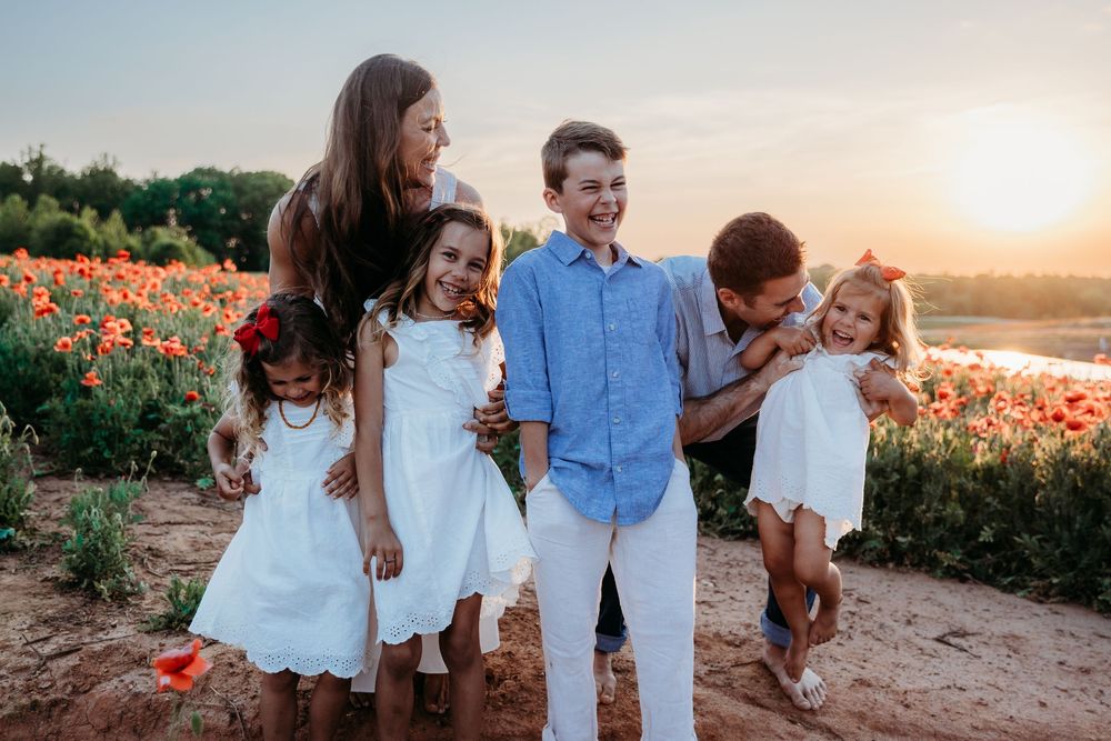 Family of five posing in a vibrant poppy field at golden sunset, children in white outfits smiling together.