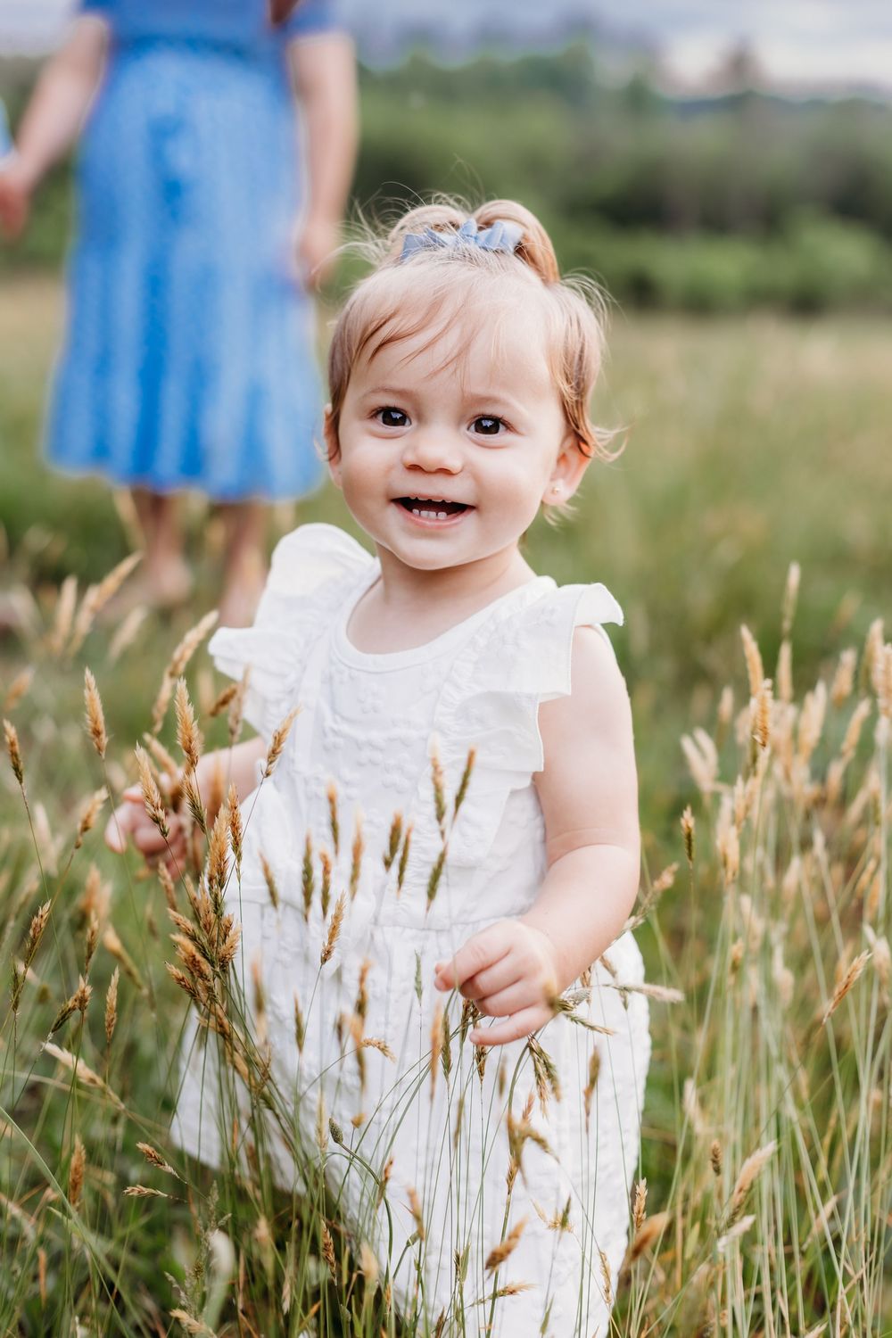 Smiling baby girl in white dress standing in a grassy field with tall wheat stalks, woman in blue dress behind her.