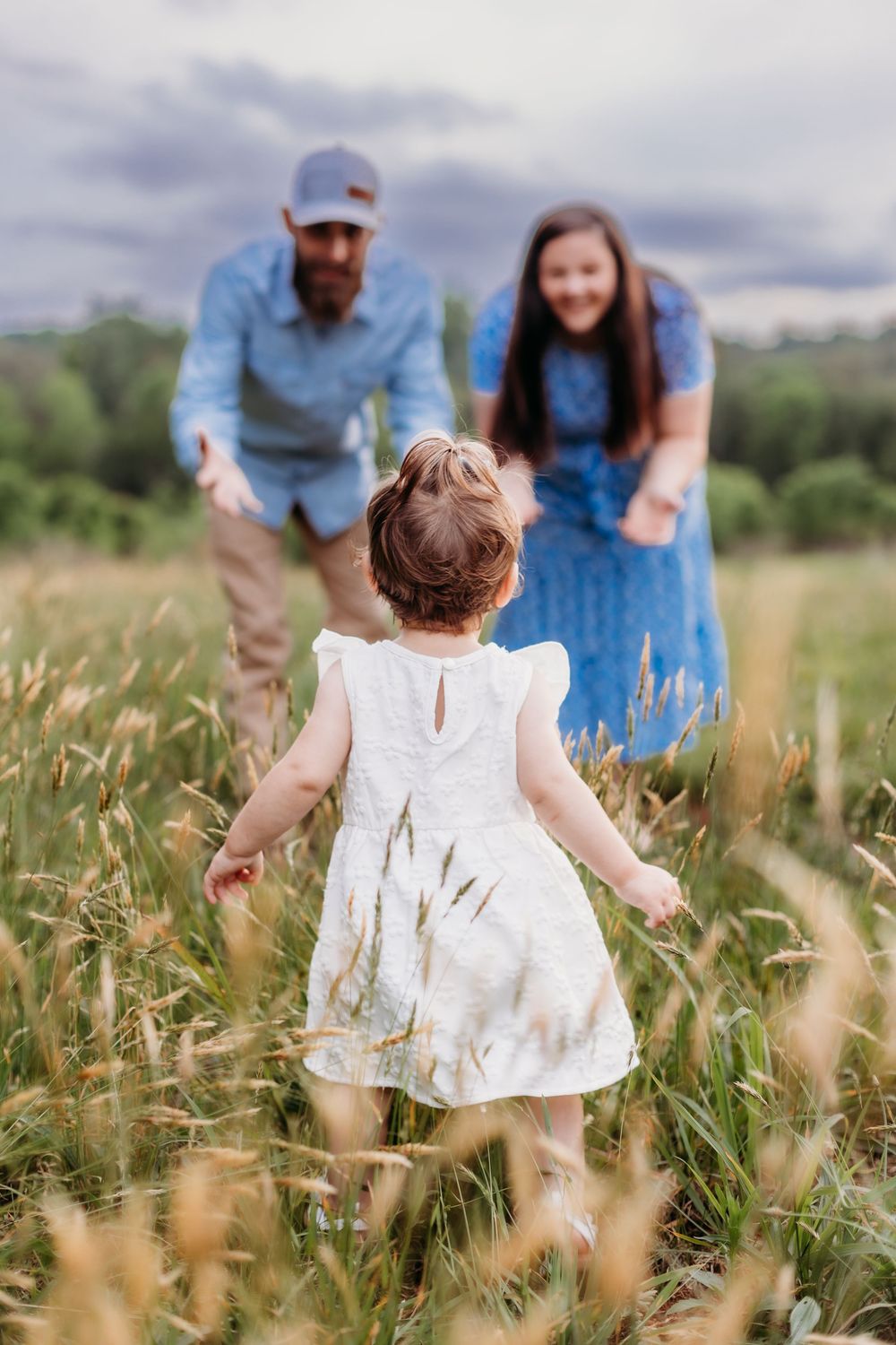 A toddler in a white dress runs through a field toward her smiling parents dressed in blue on a cloudy day.
