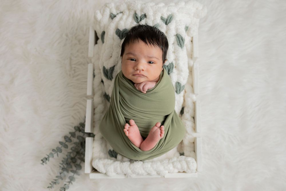 Newborn baby wrapped in soft sage green swaddle lying on white blanket with heart pattern background in his auburn hills michigan home
