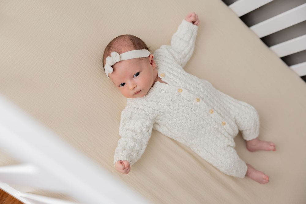Baby wearing white knitted sweater lying on white bedding in their crib in Clarkston michigan