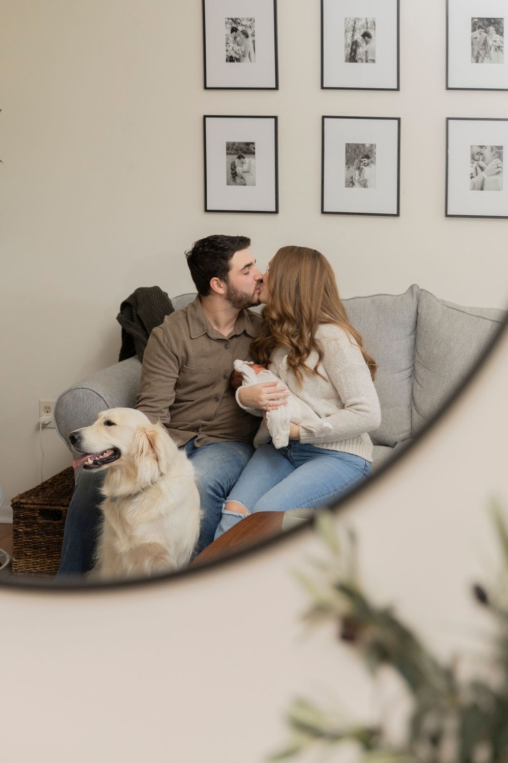 parents holding their newborn baby and kissing on their couch while petting their dog