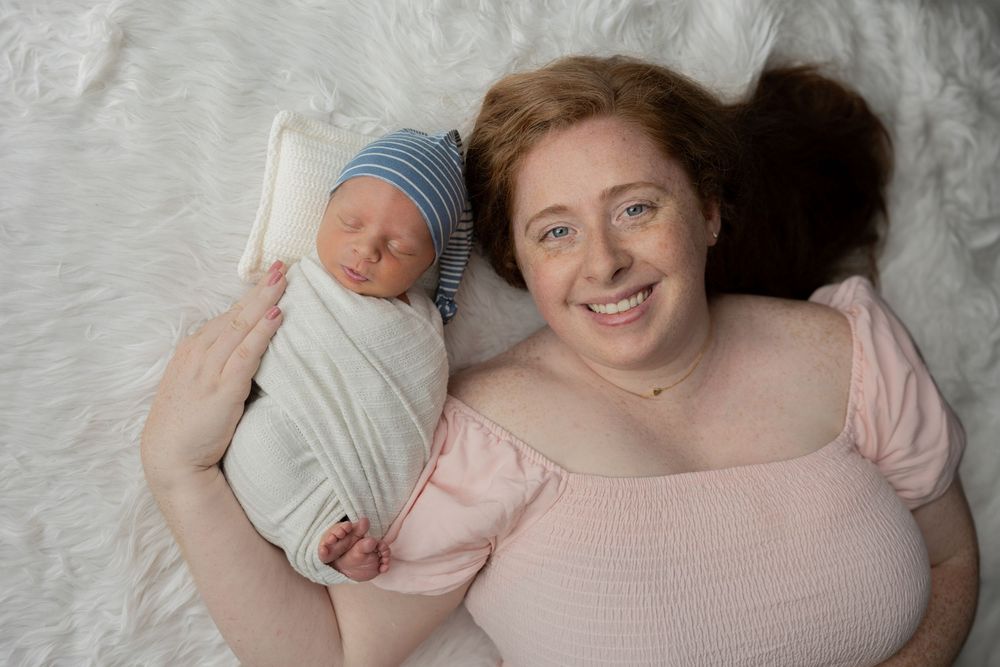 A smiling redheaded mother lies on a white fur blanket holding her swaddled newborn baby wearing a blue striped hat in her Rochester home