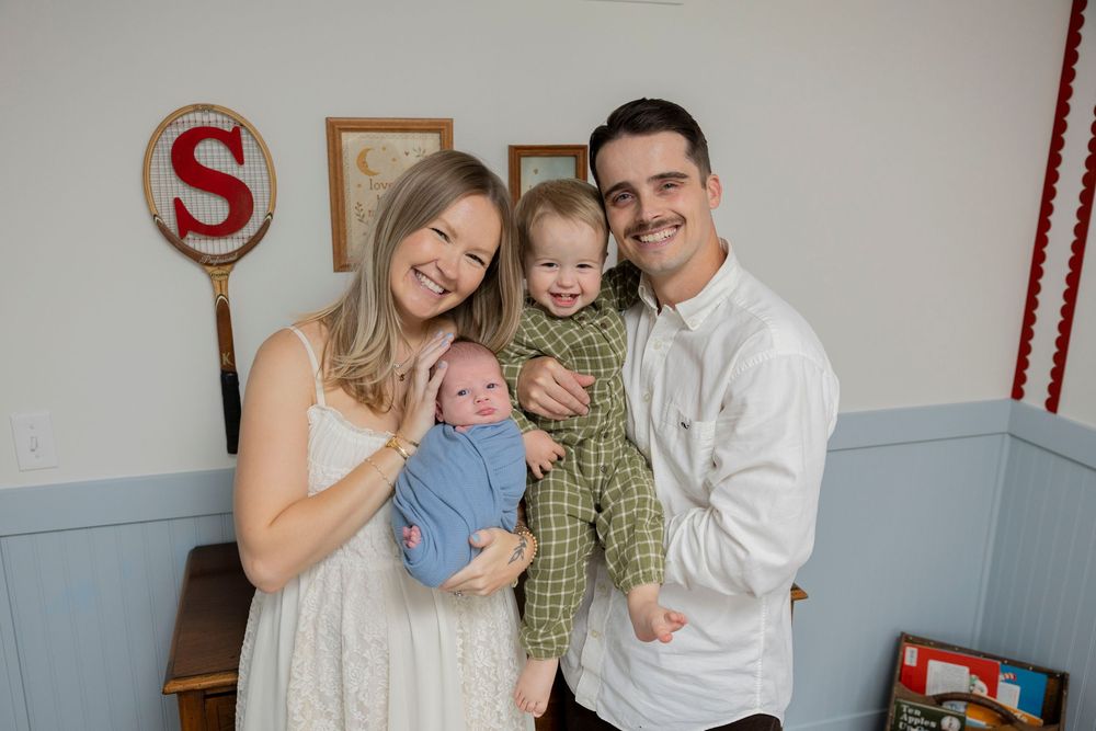 Happy family of four posing indoors in Oxford michigan, parents holding two young children, smiling in a decorated nursery room.