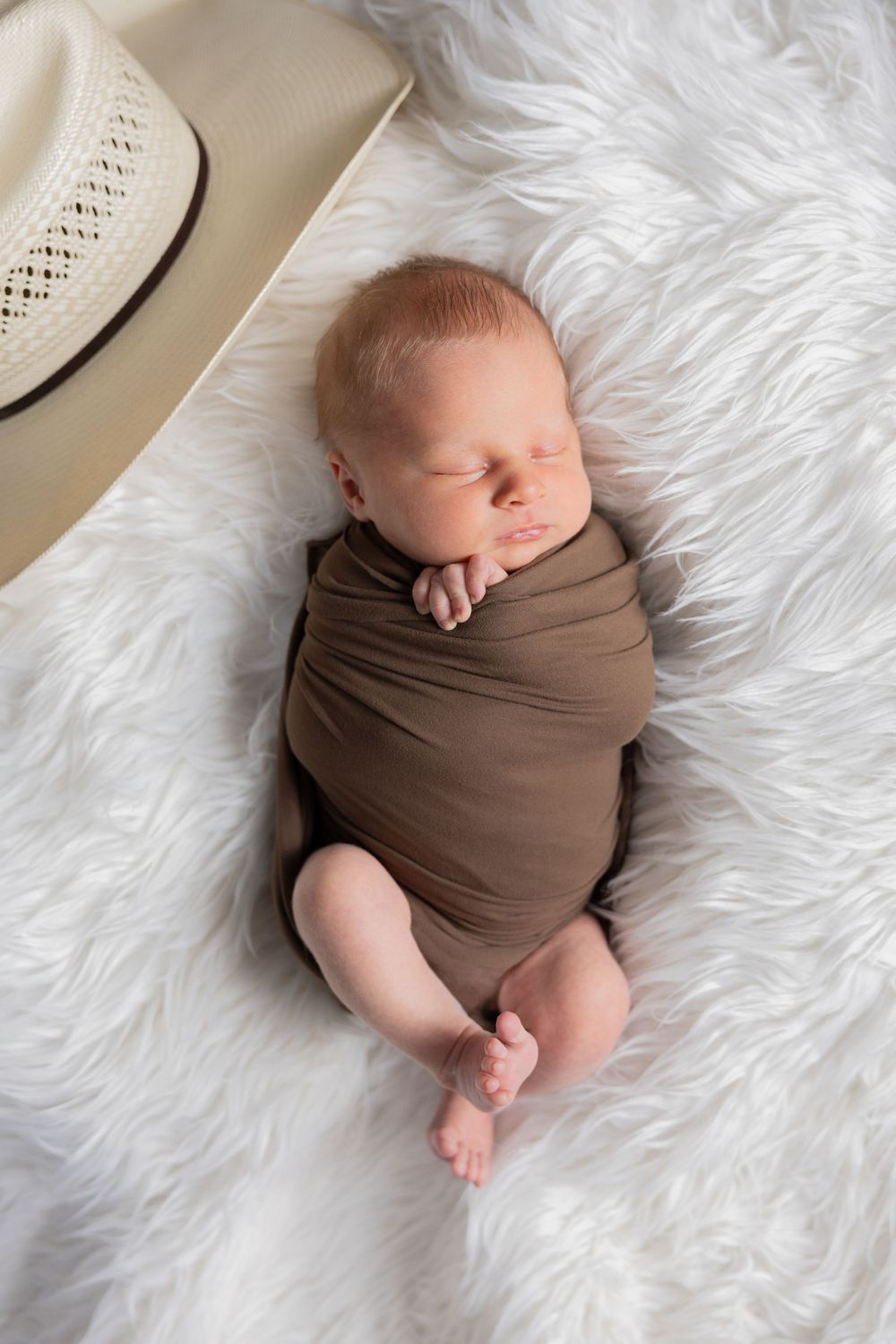 Newborn baby swaddled in brown fabric sleeping peacefully on soft white fur blanket with a cowboy hat in their Rochester hills Michigan home