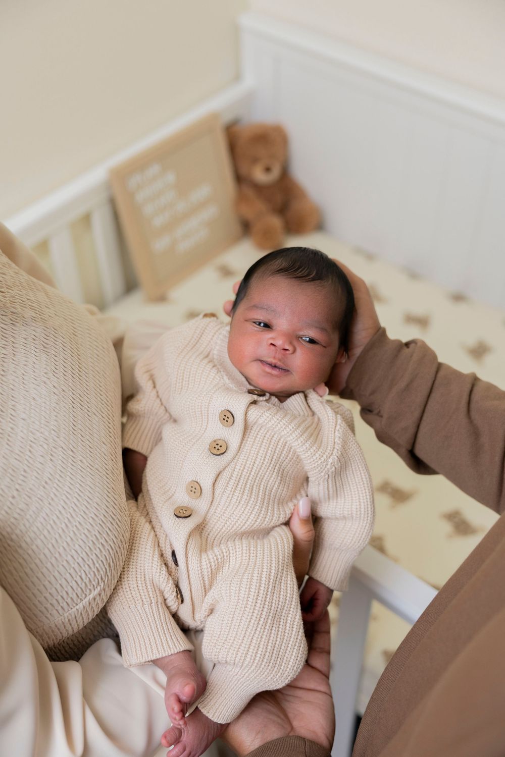 Newborn baby in cream knit cardigan held by parent near white crib with teddy bear and bear-print bedding in metro Detroit michigan