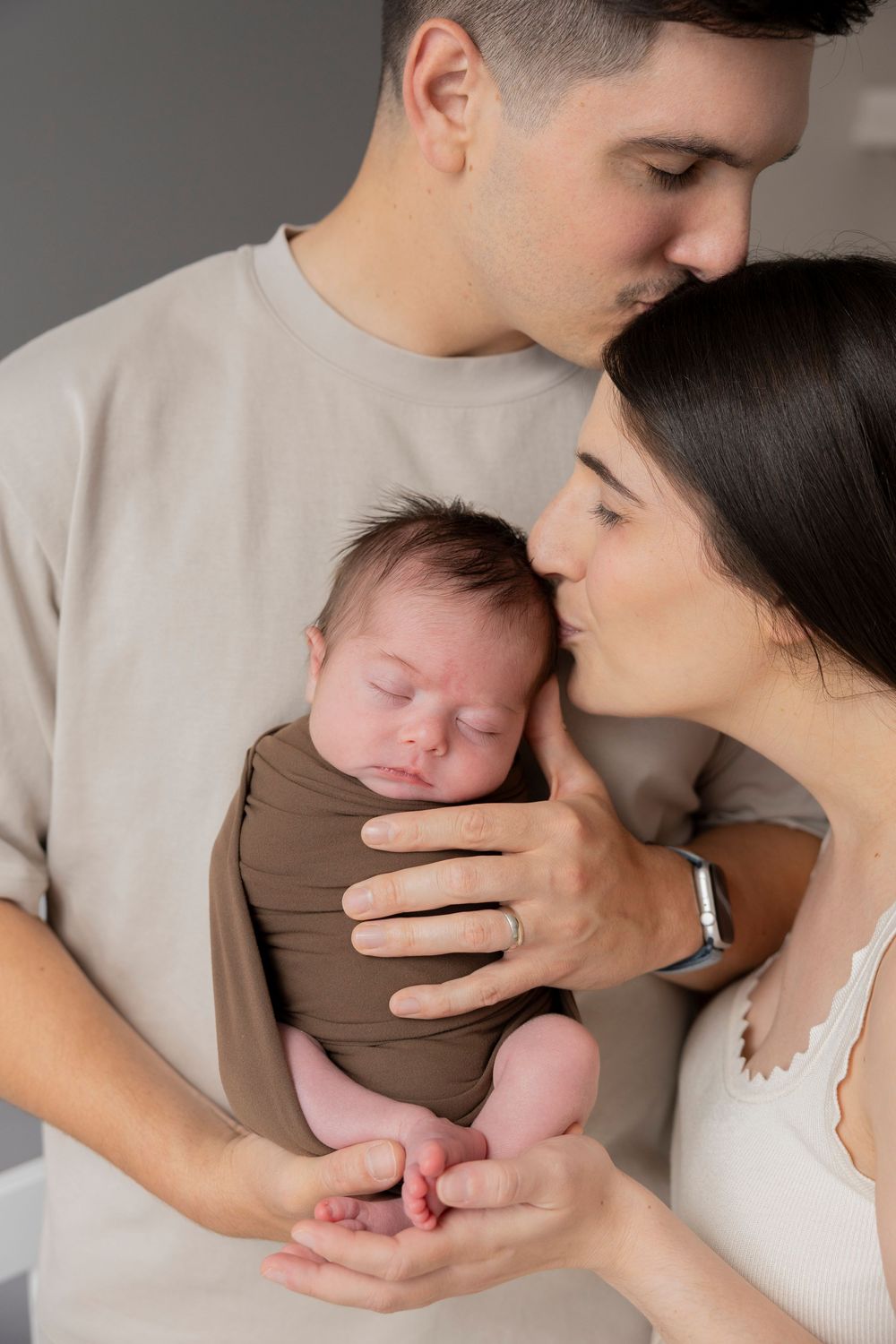 A tender moment as parents cradle and embrace their sleeping newborn baby wrapped in brown swaddle in their Oakland county Michigan home