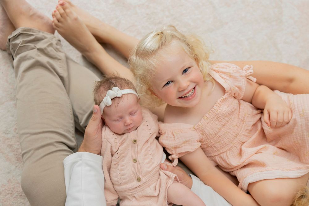 sisters wearing a peach-colored outfit lies on their parents lap and smile for a newborn photo in their Rochester hills michigan home