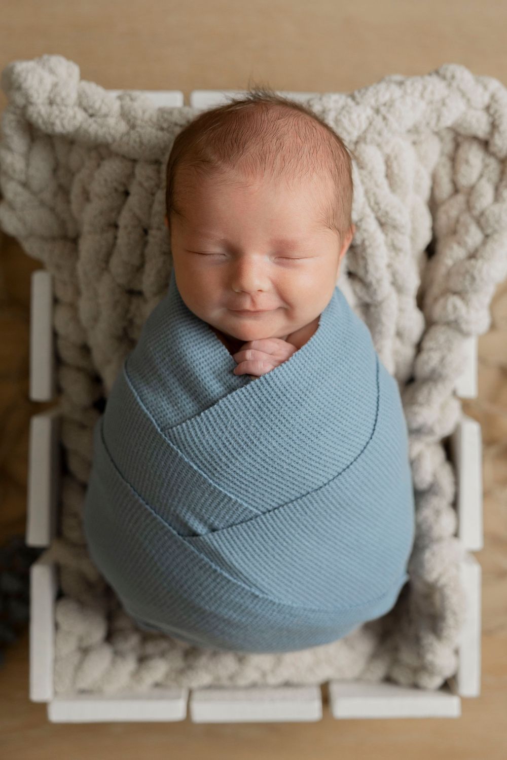 Sleeping newborn baby swaddled in a light blue wrap, resting in a white wooden crate with a cozy knit blanket.