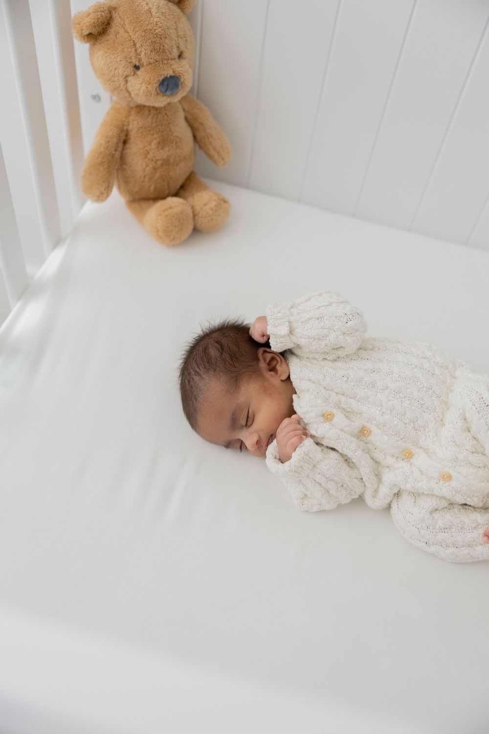 A sleeping newborn baby in white knit clothing lies on white bedding with a brown teddy bear in the background in his Detroit home