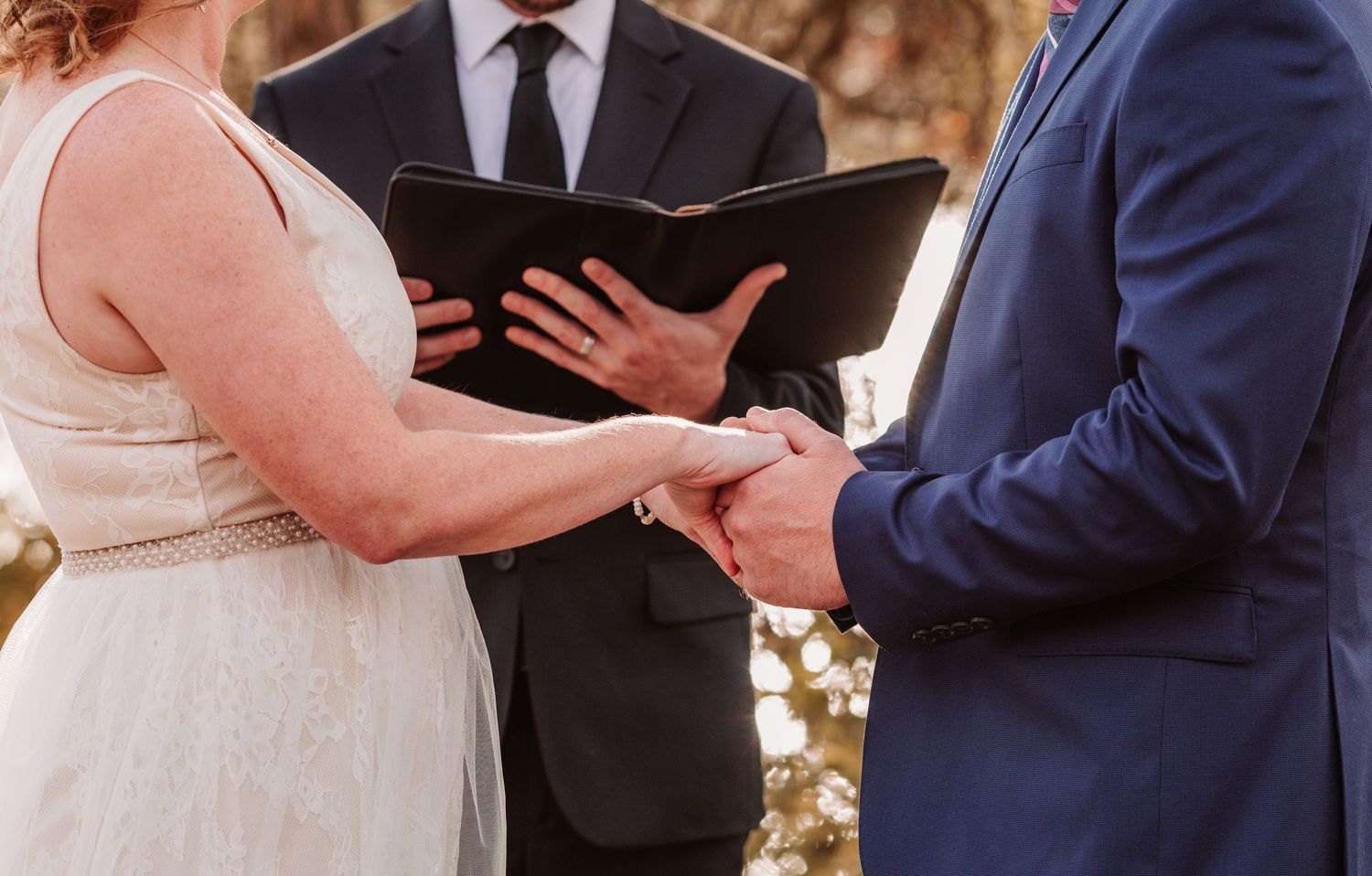 Bride and groom exchange vows holding hands while officiant reads from a black book at outdoor wedding.