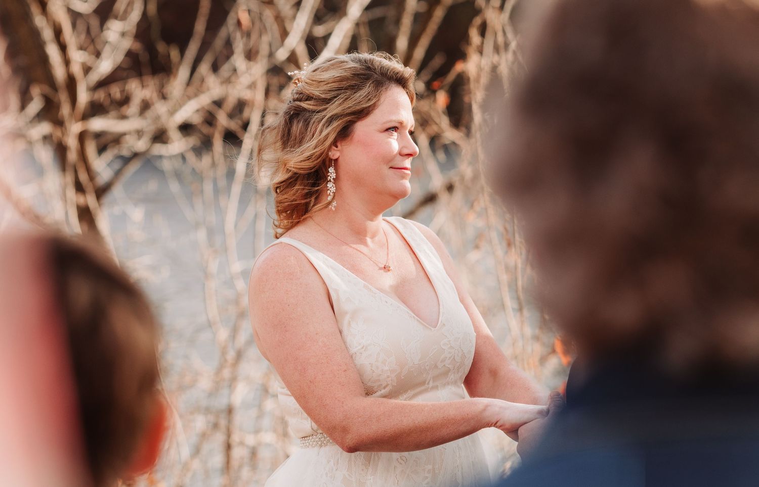 Smiling bride in sleeveless lace wedding gown stands at outdoor autumn wedding ceremony.