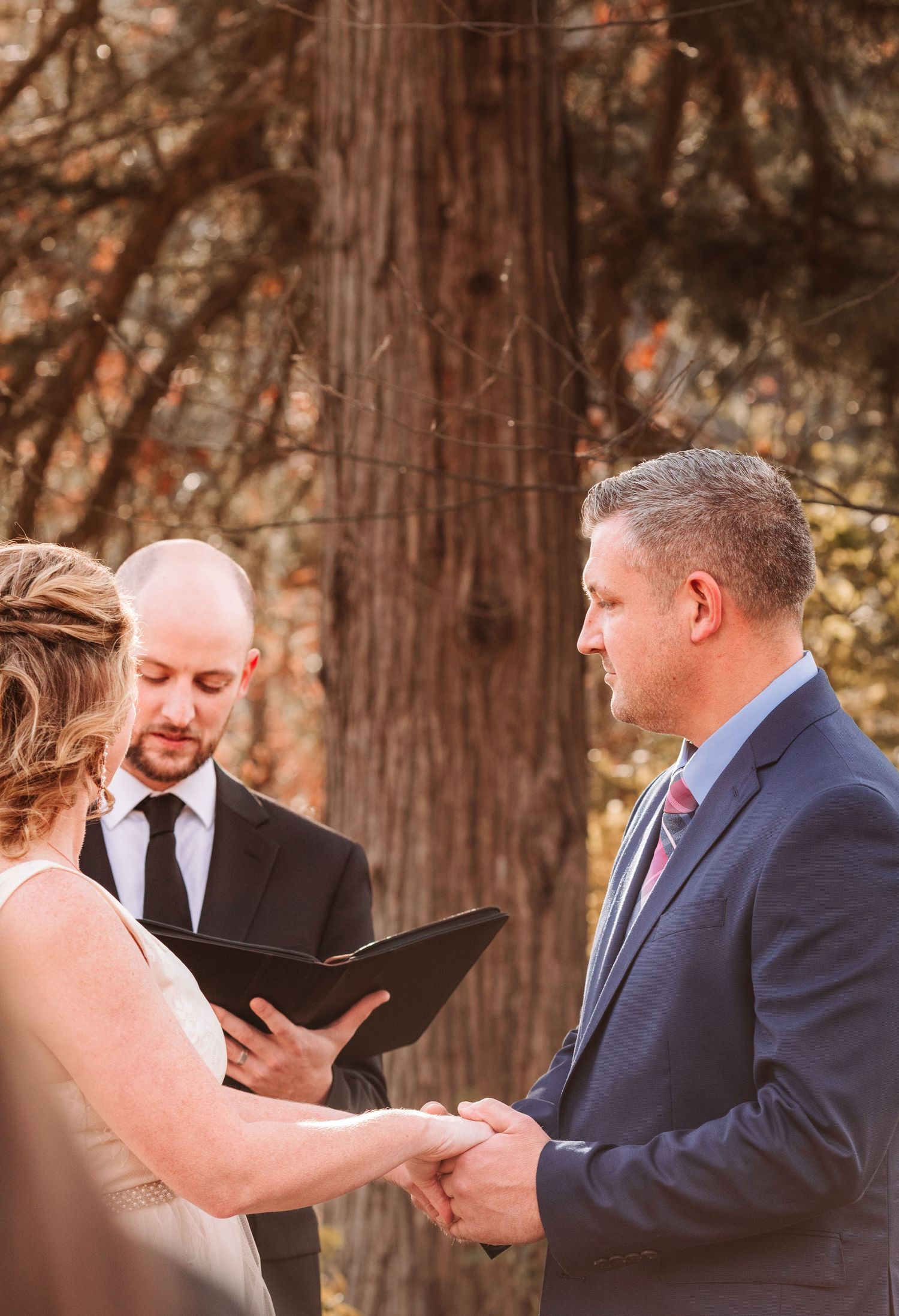 Groom in navy suit holds brides hands during outdoor wedding ceremony with officiant reading vows.