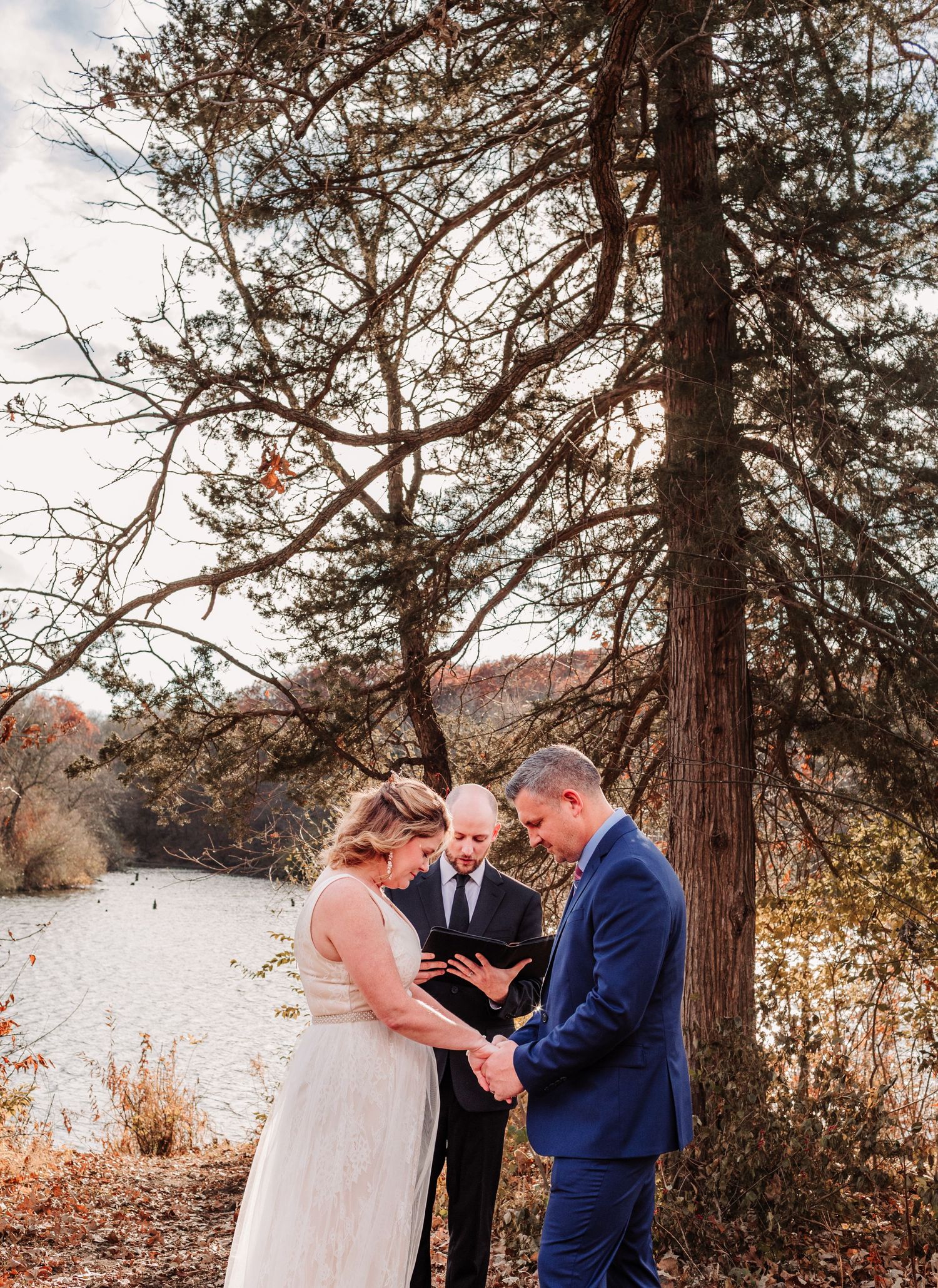 Bride and groom hold hands during intimate outdoor fall wedding ceremony beside a serene lake.