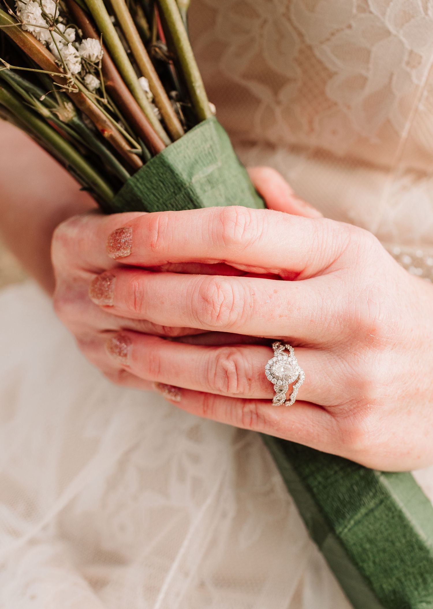Close-up of brides hand holding bouquet wrapped in green ribbon, featuring a halo diamond engagement ring.