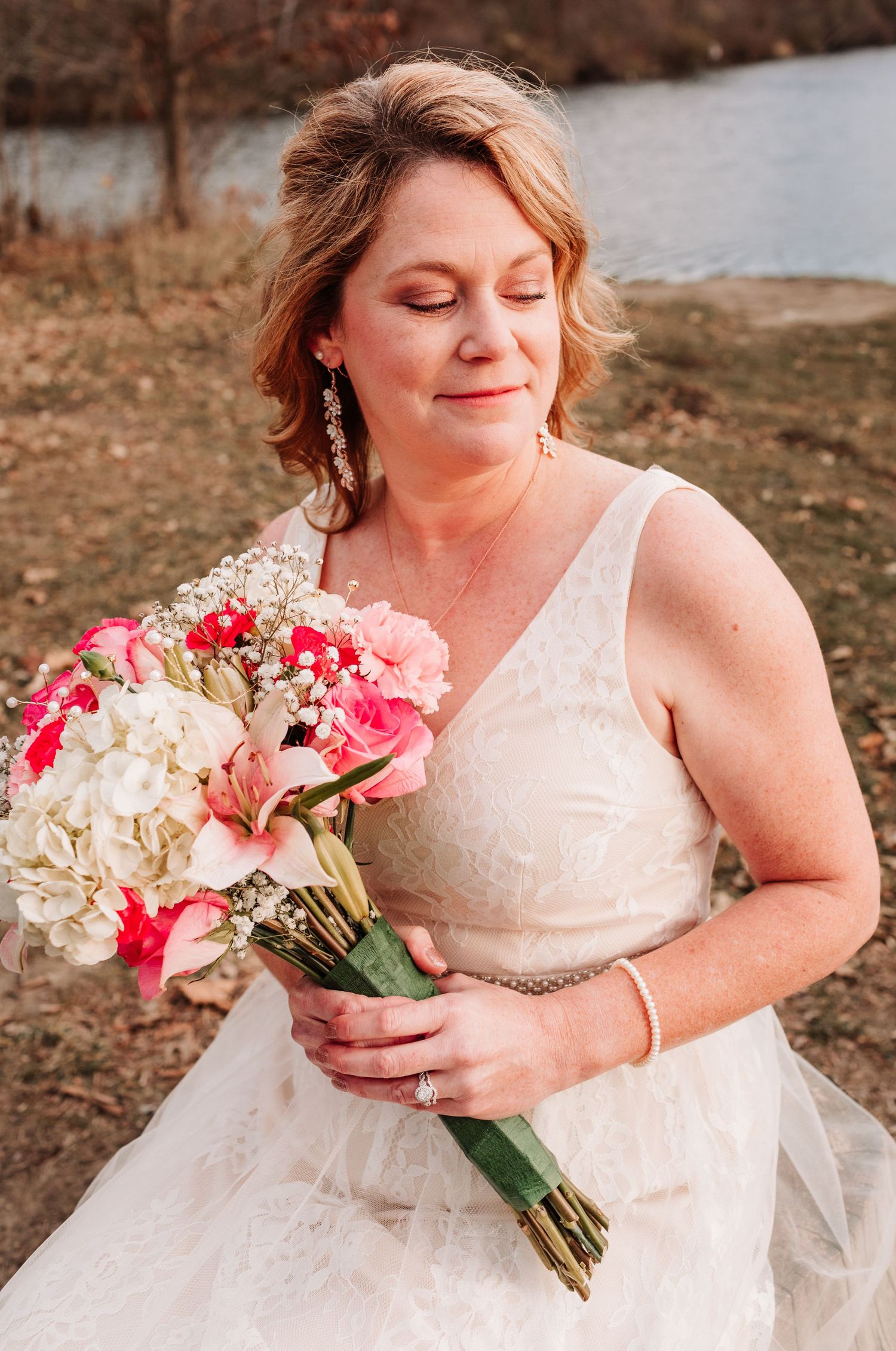 Bride in lace wedding gown holds colorful pink and white bouquet with eyes closed near a lake.