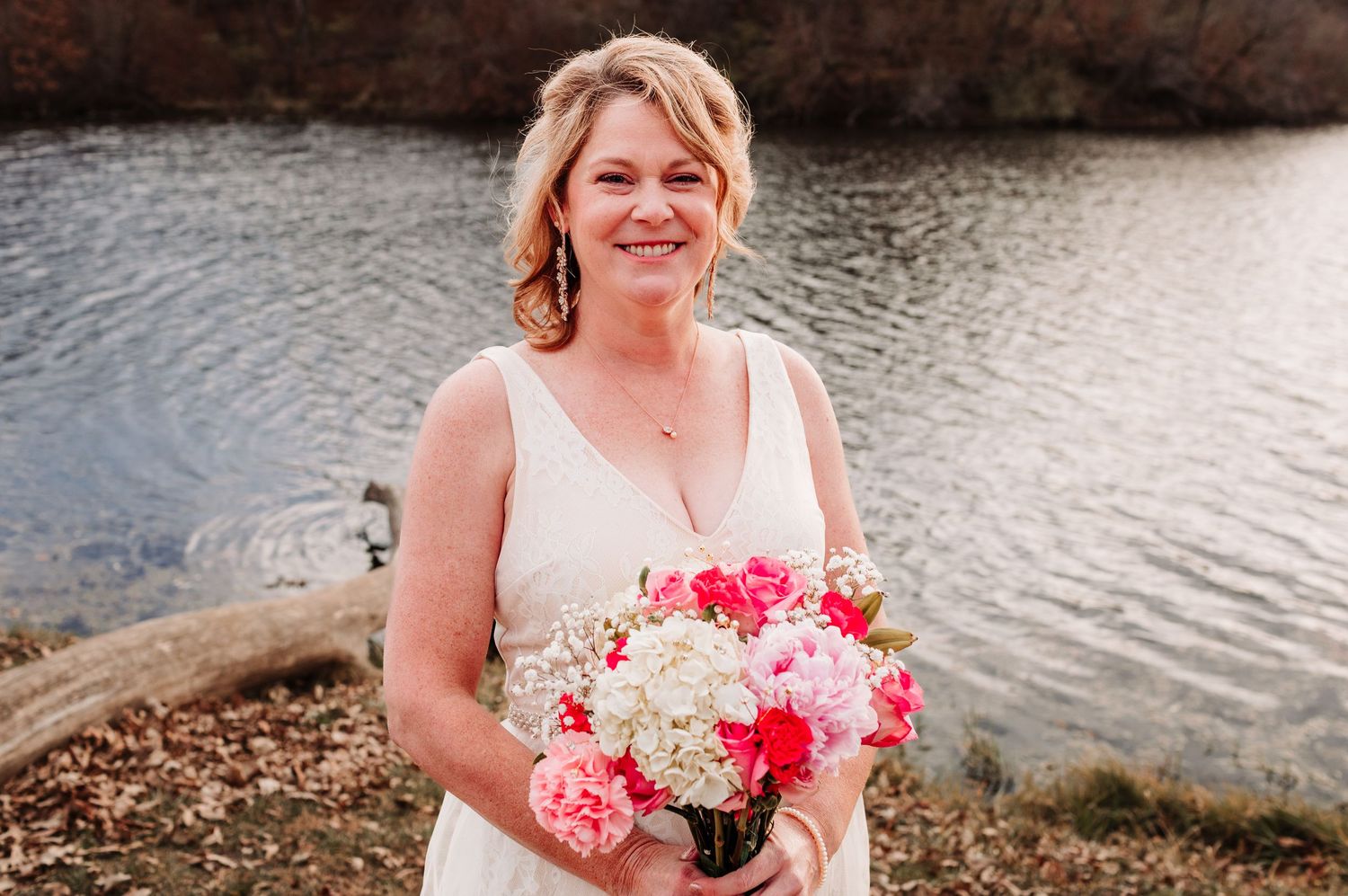 Smiling bride in white dress holding pink and white floral bouquet standing by a serene lake.