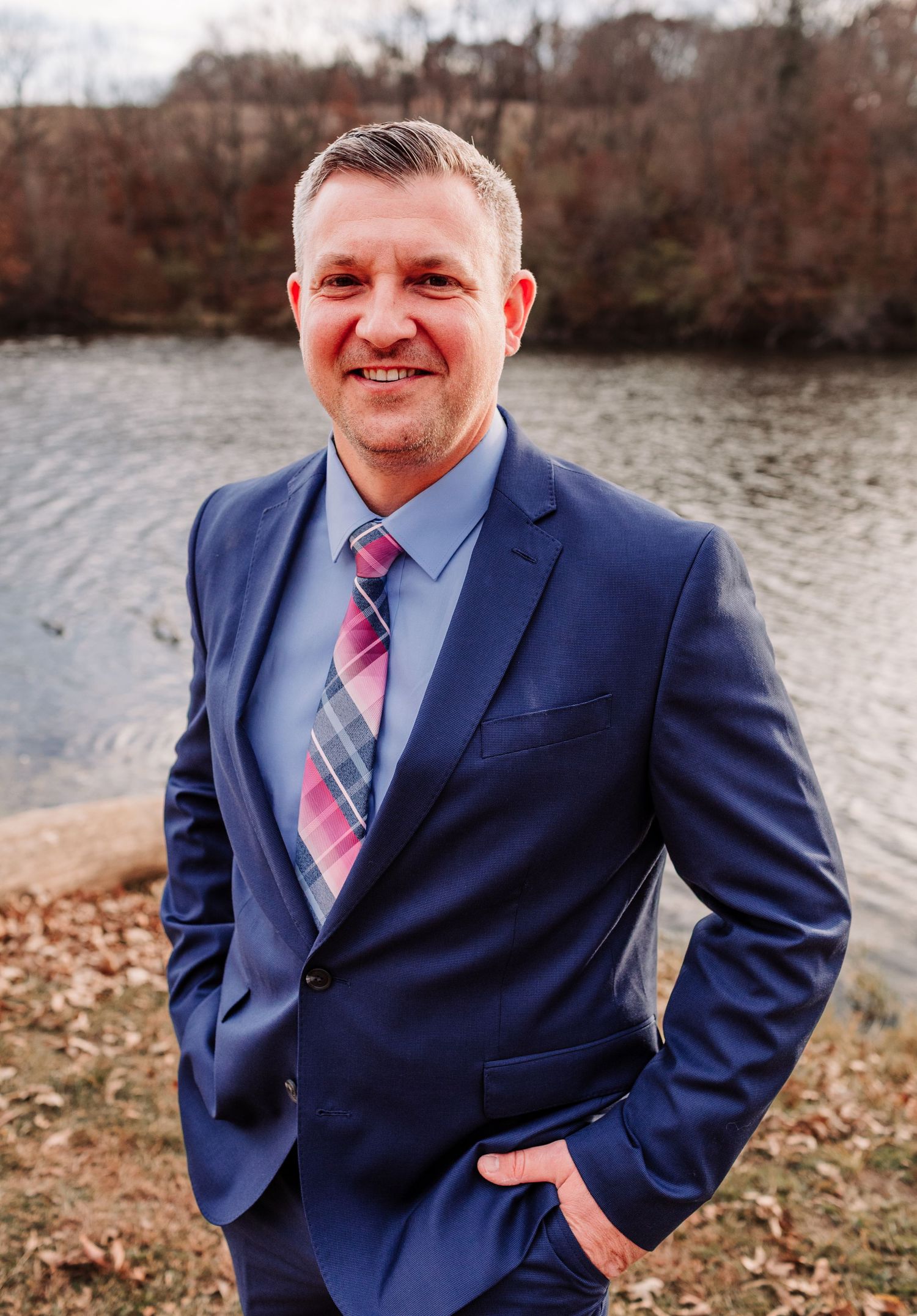 Groom in navy suit and pink plaid tie smiling confidently by a lakeside backdrop.