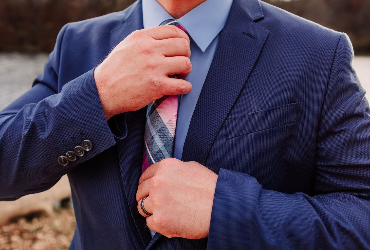 Groom in navy blue suit adjusting his pink plaid tie with wedding ring visible on finger.