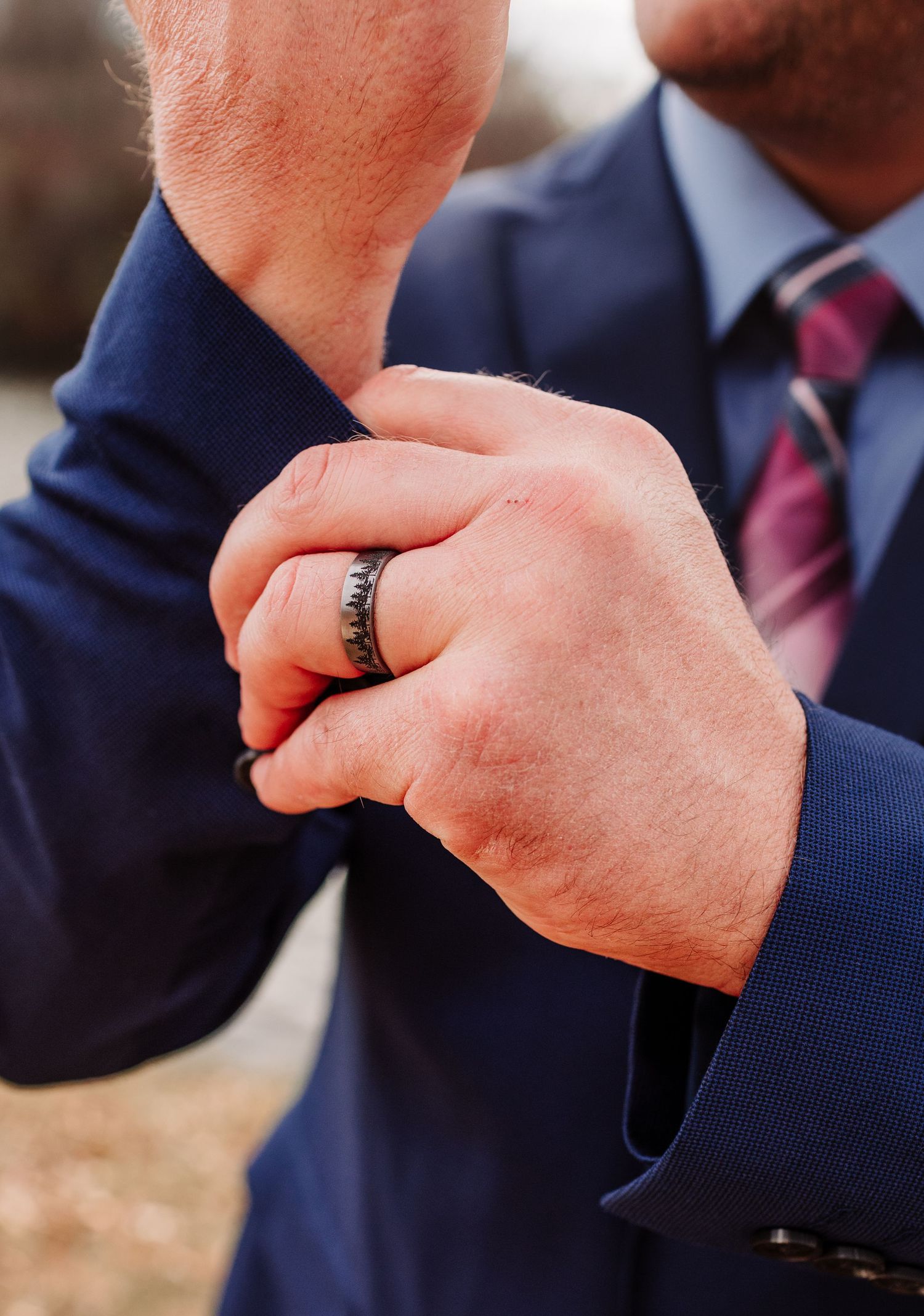 Close-up of groom adjusting navy suit sleeve with black wedding band on finger.