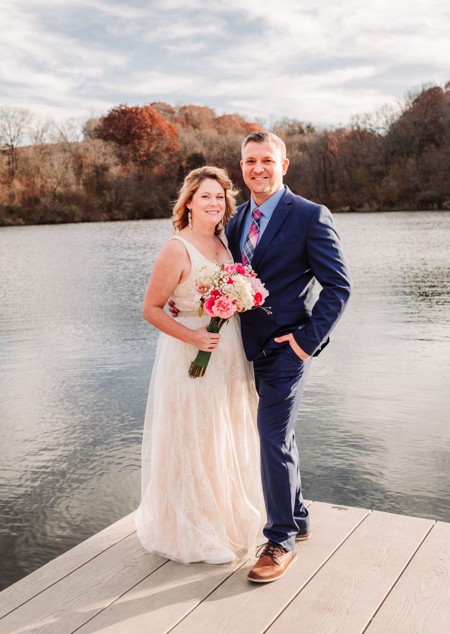 Bride and groom posing on a dock by a lake, she holds a pink bouquet in fall setting.