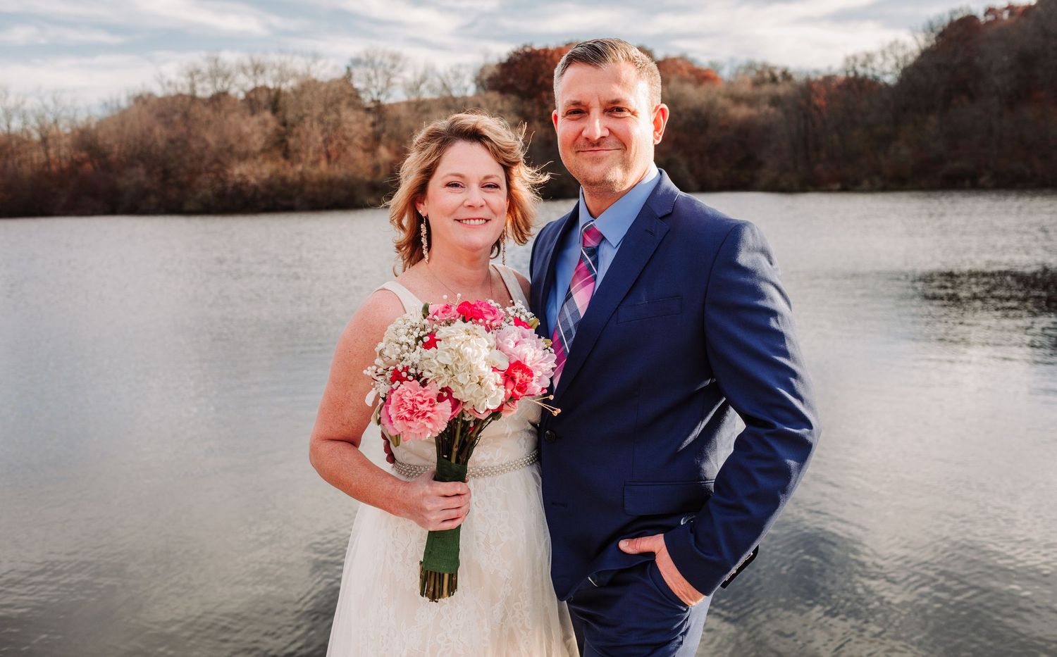 Bride and groom smiling together by a lake, she holds pink flowers in autumn scenery.