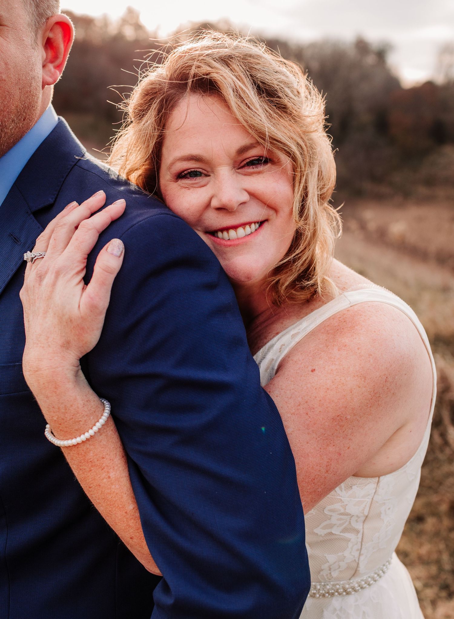 Radiant bride in white dress hugging groom in navy suit, smiling warmly in golden light.