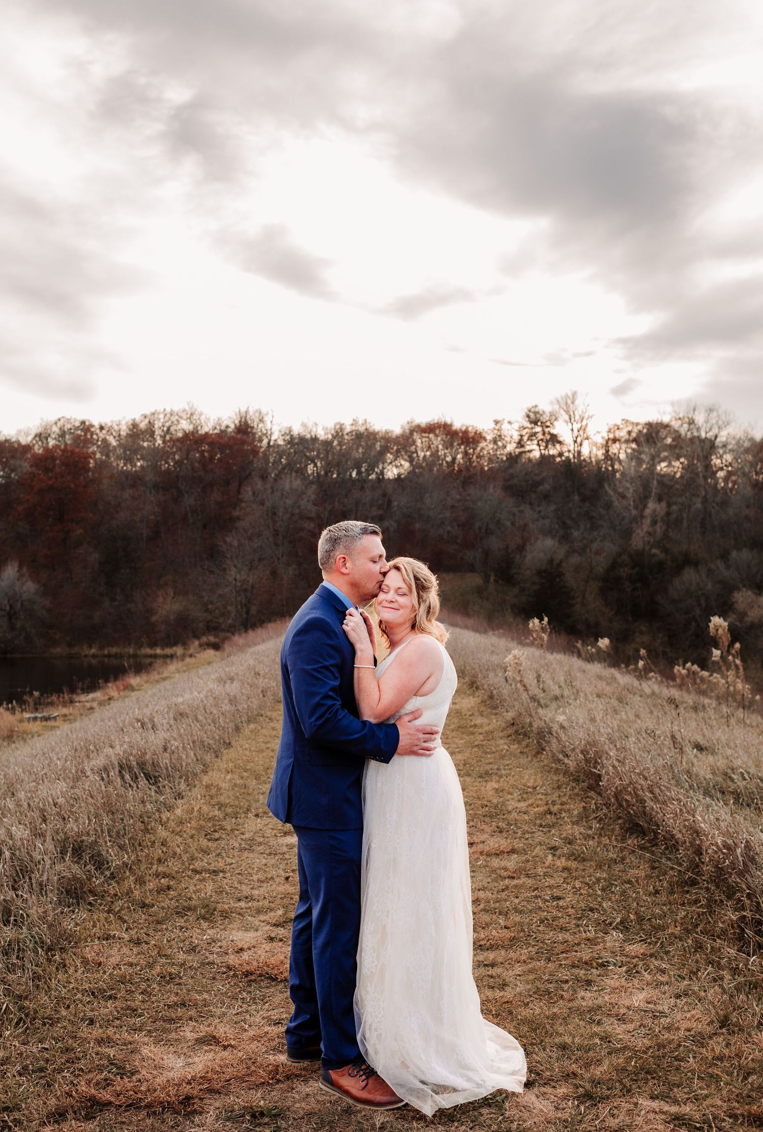 Bride and groom sharing a kiss on a scenic rural path surrounded by autumn foliage.