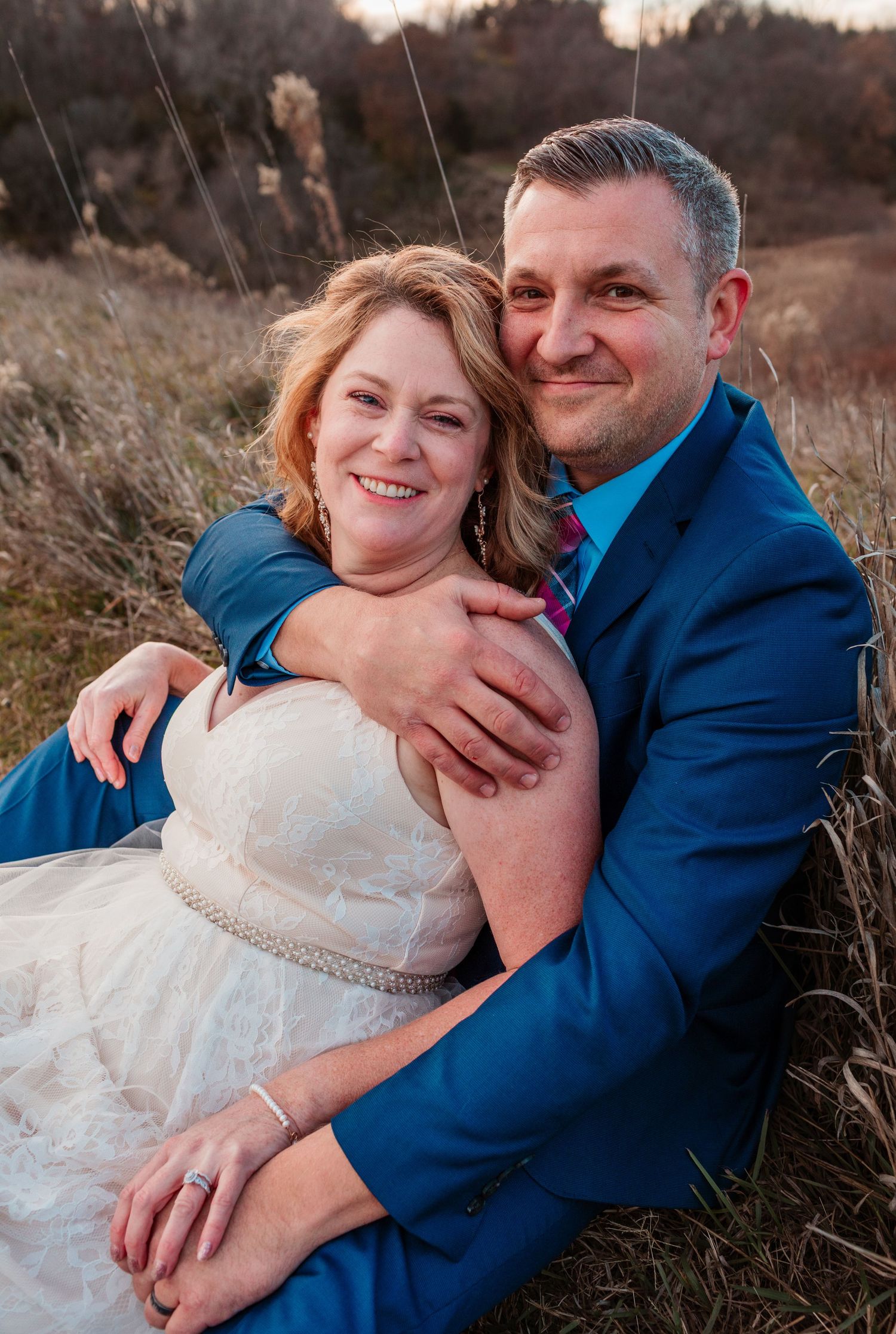 Bride and groom sitting in a golden field, smiling warmly as groom embraces bride in white wedding dress.