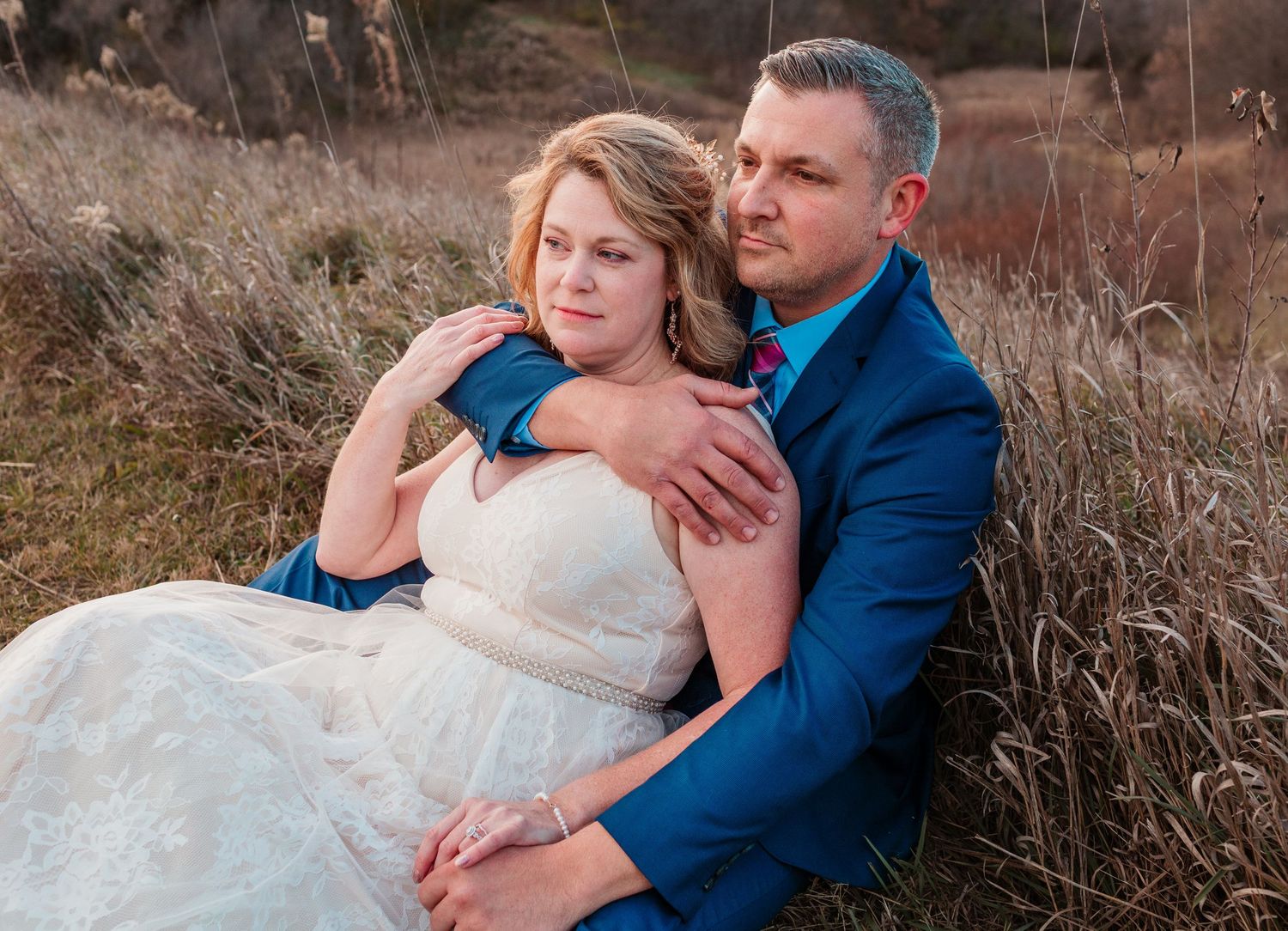 Bride and groom sitting together in a golden field, groom embracing bride from behind.