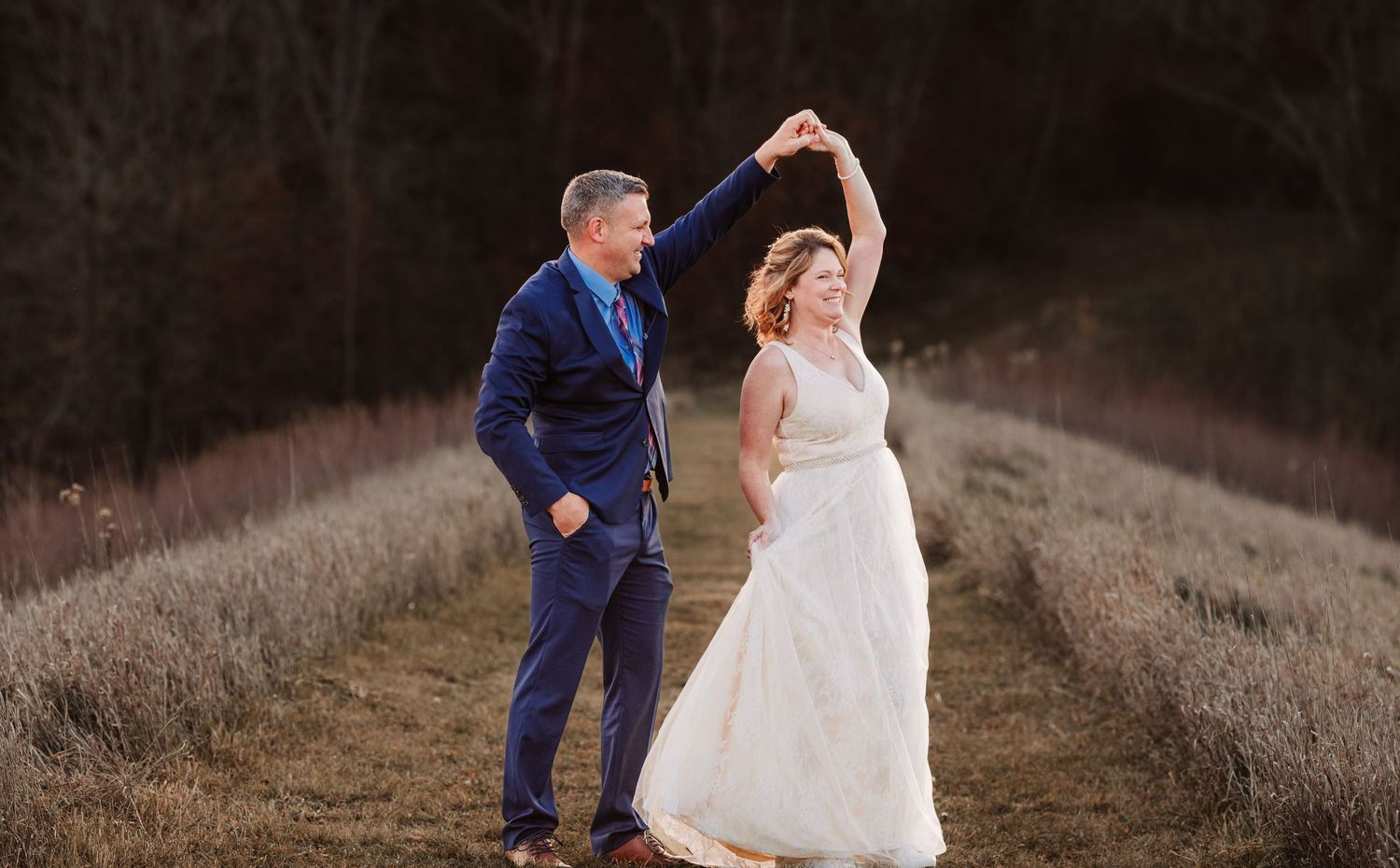 Bride and groom dancing outdoors in a field, groom spinning bride in white wedding gown at sunset.