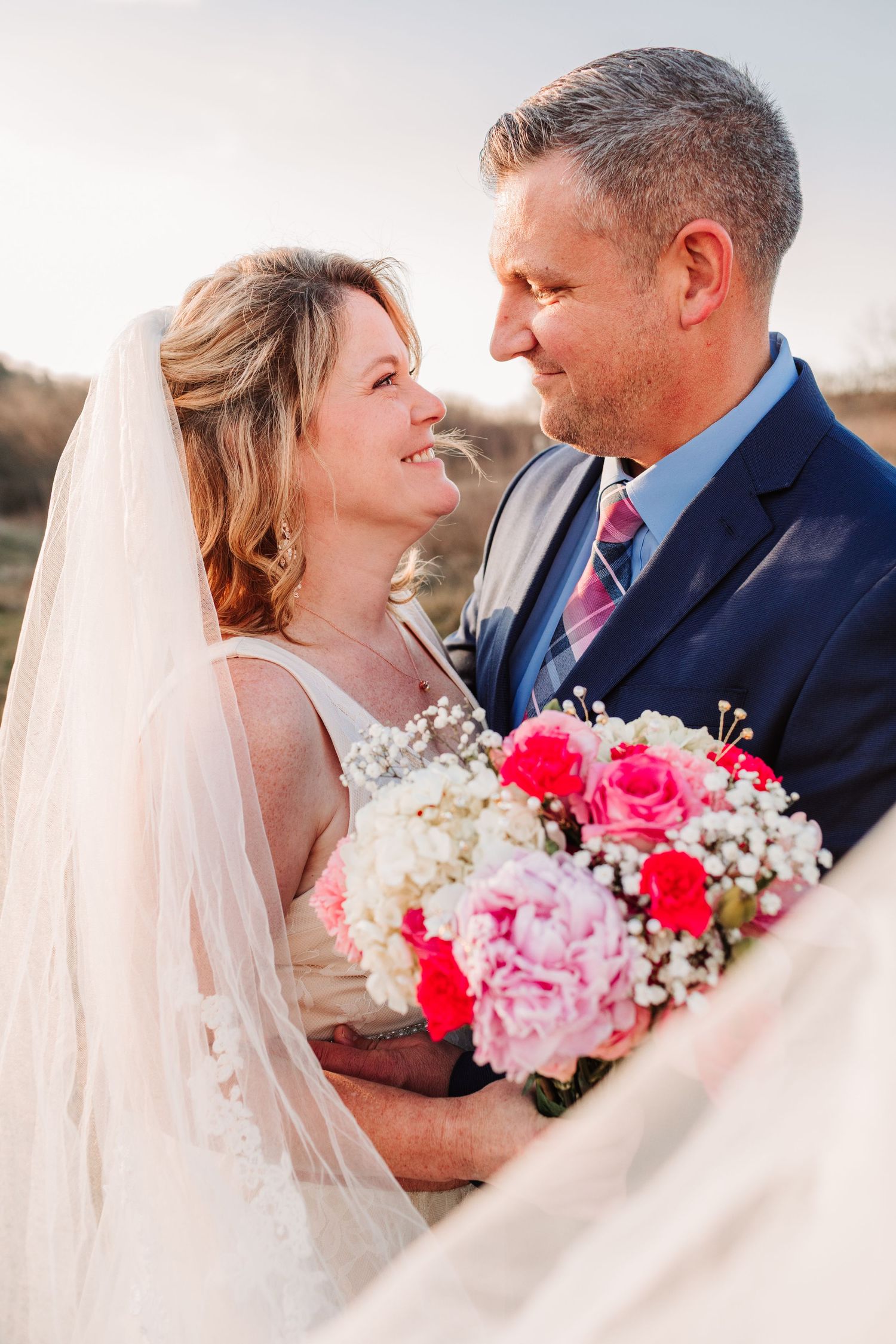 Bride and groom gazing at each other, bride holding pink and white bouquet with veil flowing in breeze.