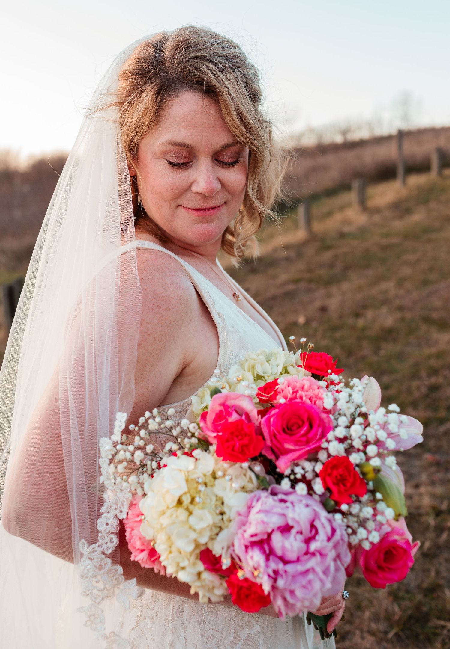 Bride in white veil looking down at vibrant pink and white wedding bouquet in outdoor setting.