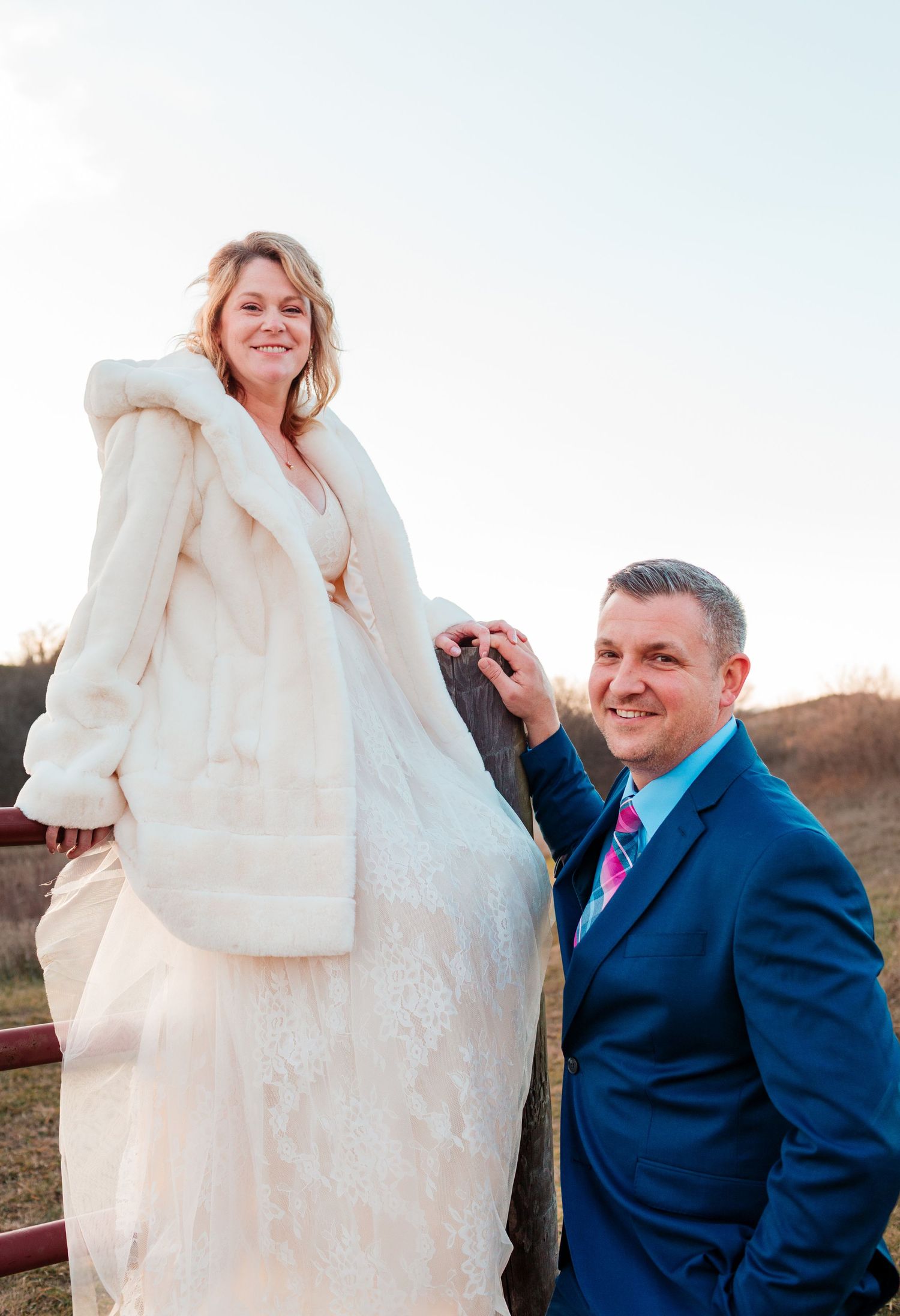 Bride in white fur coat standing elevated on fence while smiling groom in blue suit assists her.