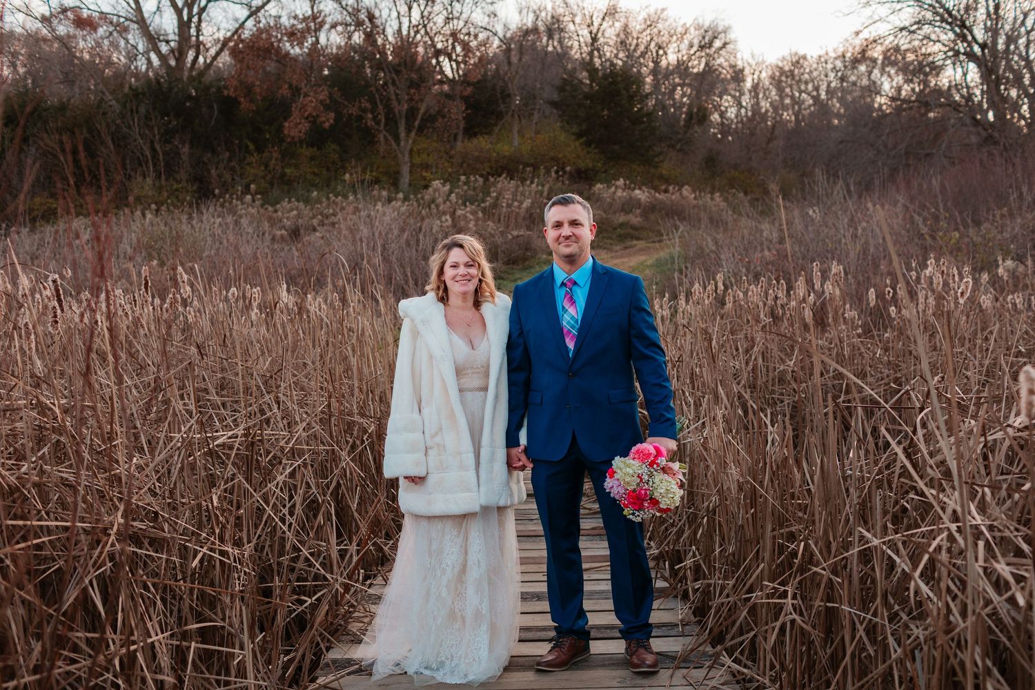 Bride and groom holding hands on wooden boardwalk surrounded by tall golden reeds at sunset.