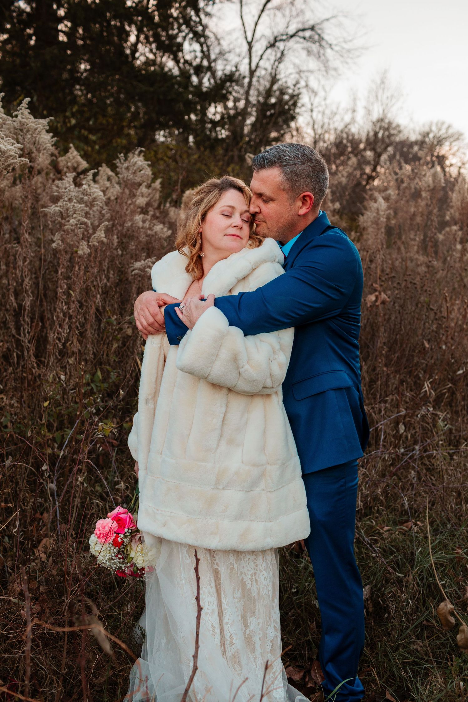 Groom in blue suit tenderly embracing bride in white fur coat among tall dried grasses outdoors.