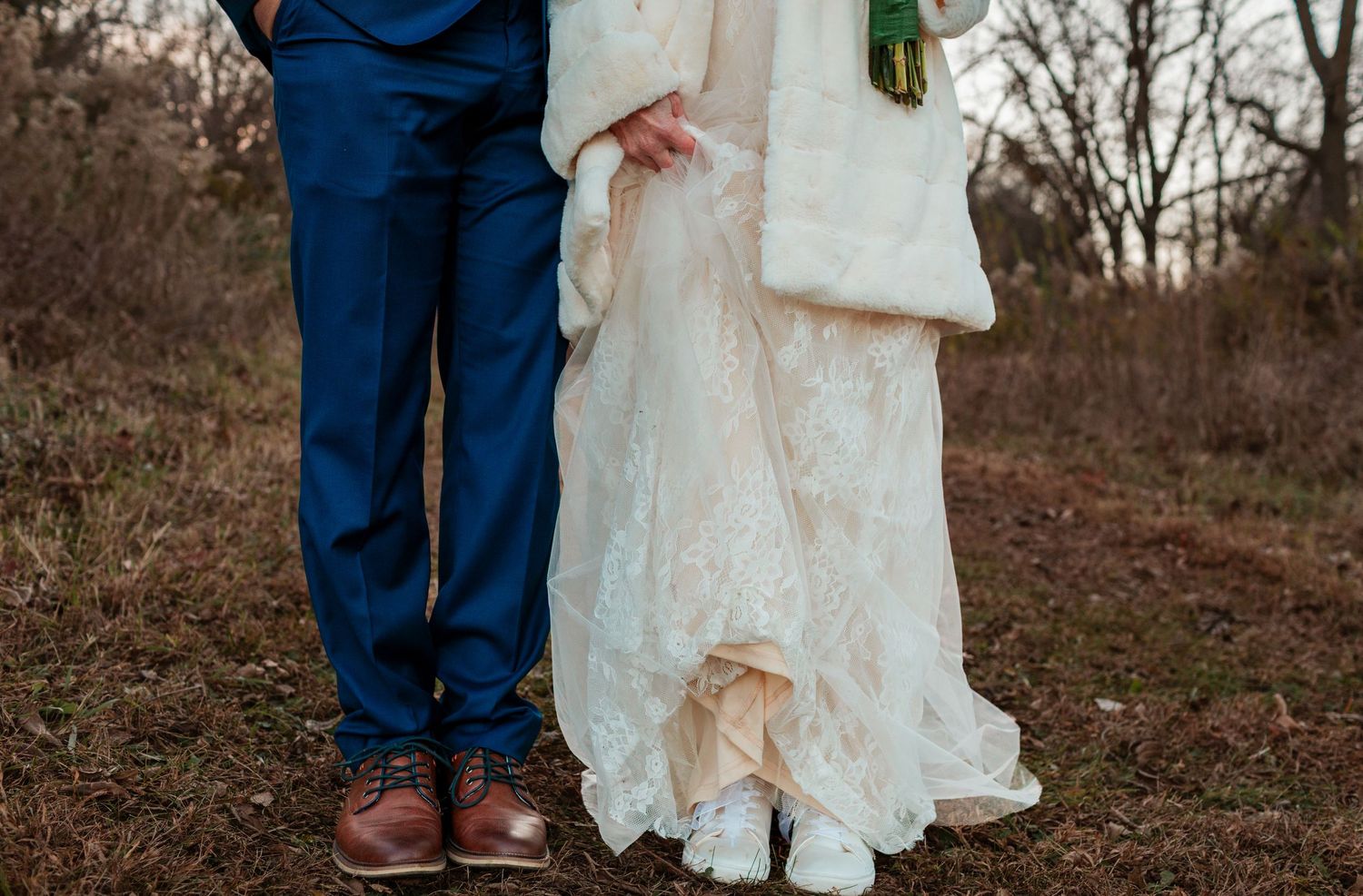 Close-up of bride in white lace gown and groom in blue suit showing their feet and attire outdoors.