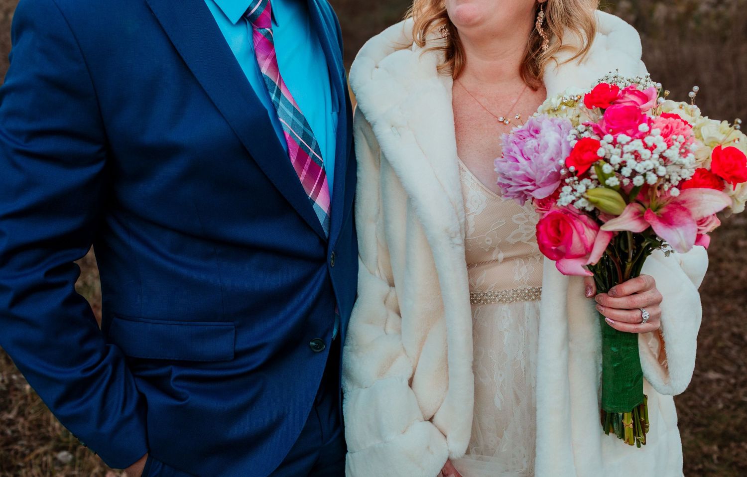 Close-up of bride in white fur coat holding colorful pink wedding bouquet beside groom in blue suit.
