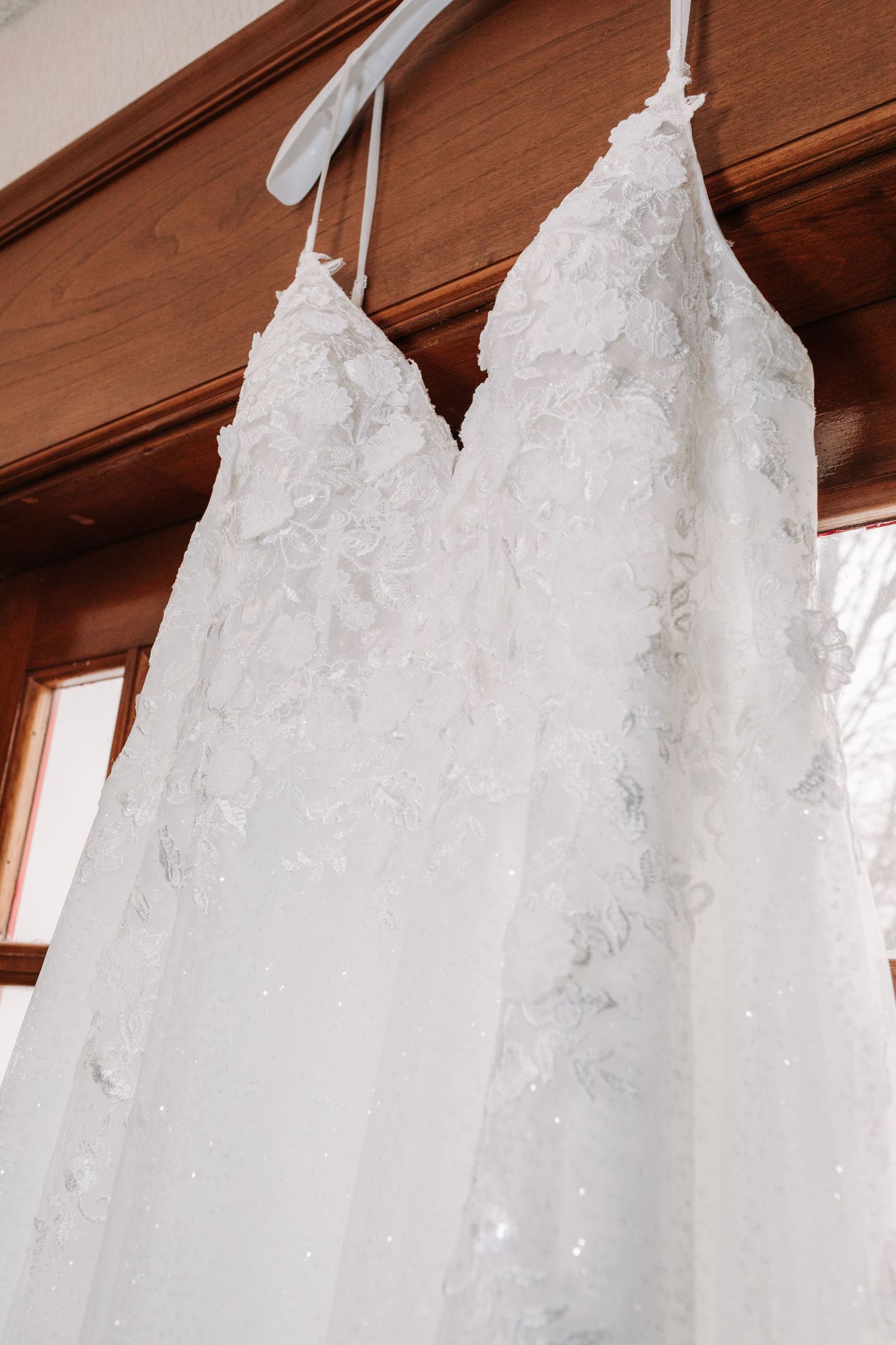 Close-up of white floral lace A-line wedding dress hanging near window.