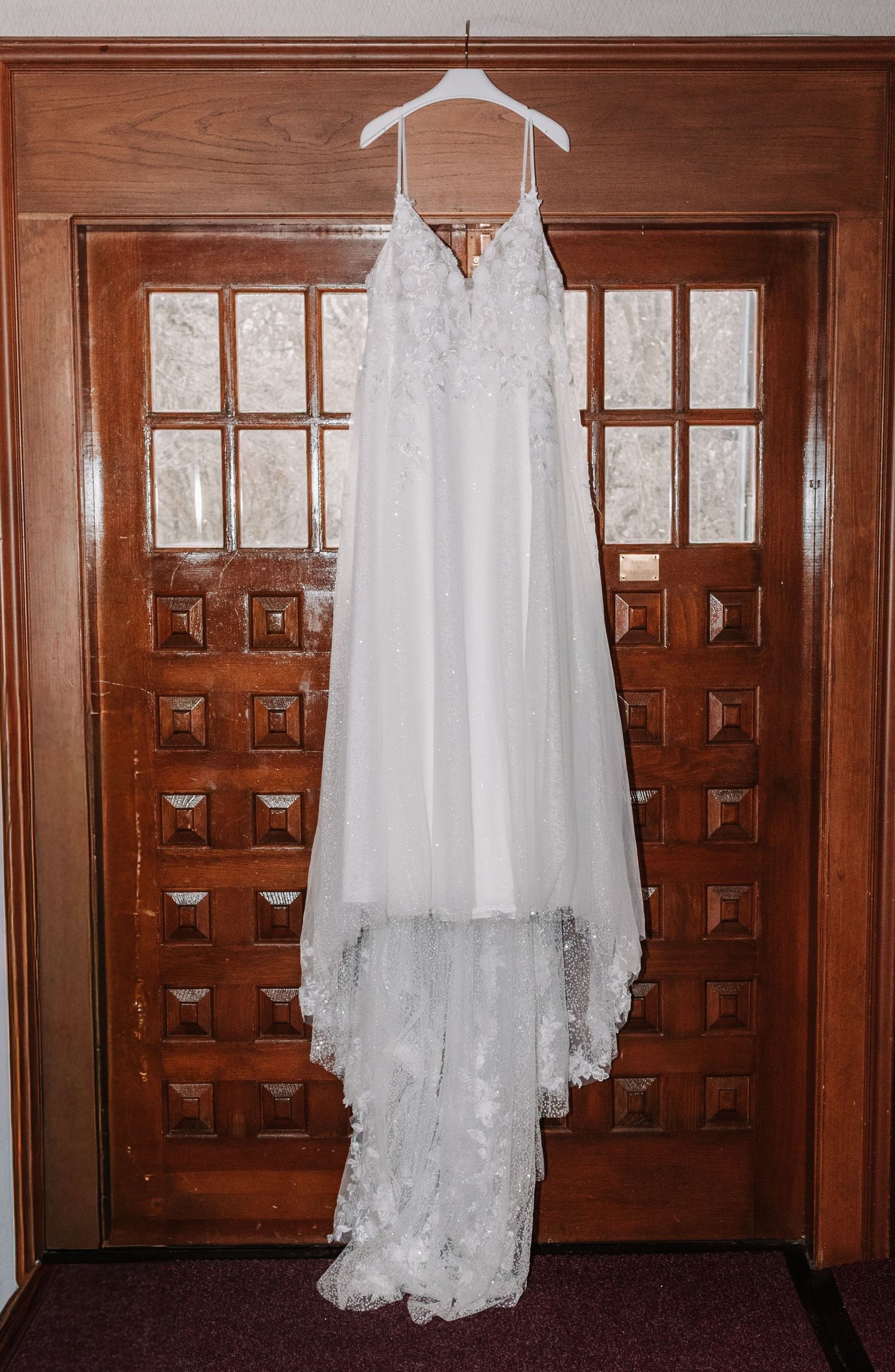 Elegant white lace wedding dress hanging on wooden door before ceremony.