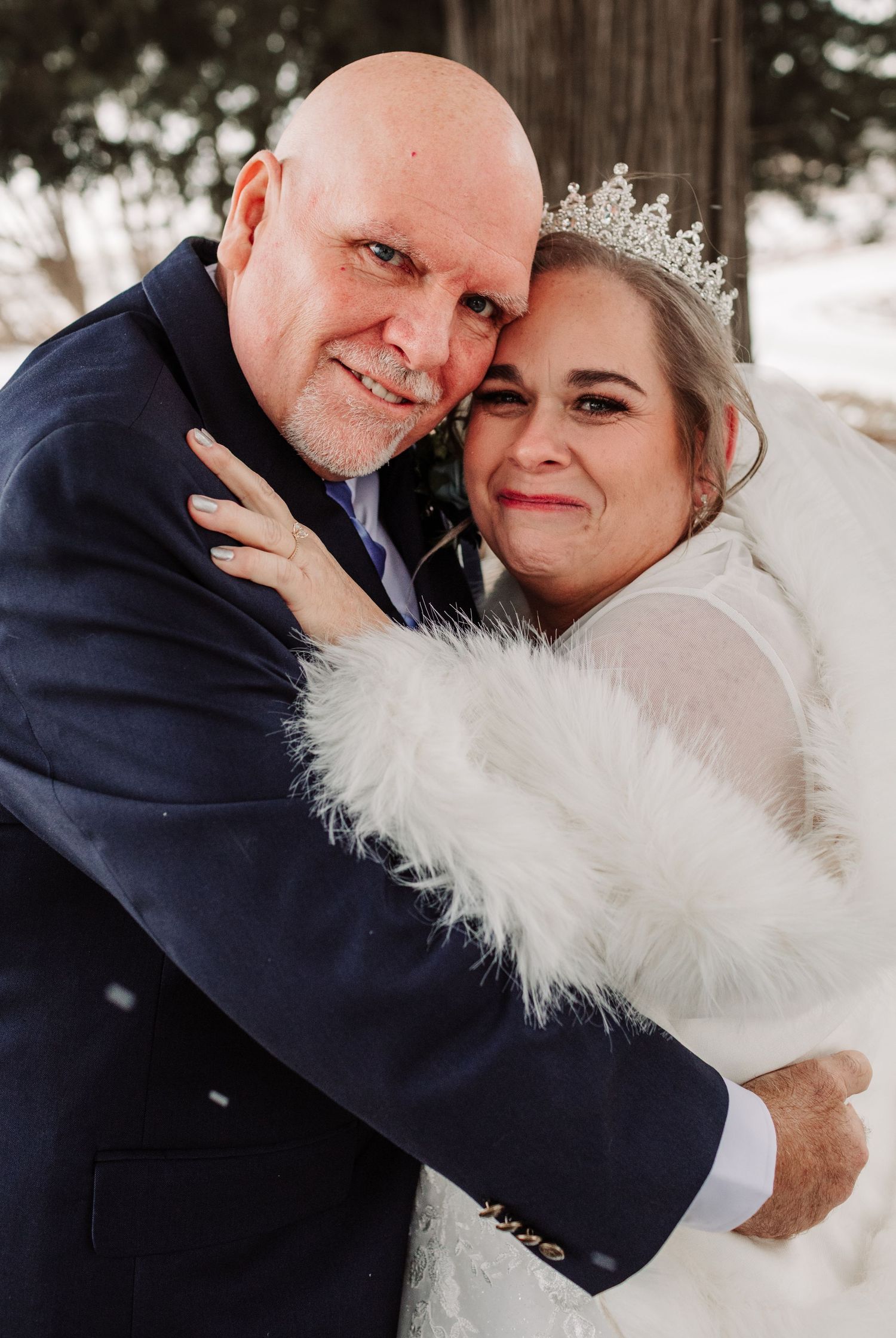 Bride wearing tiara and white feather wrap embracing bald groom in blue suit in snowy outdoor setting.