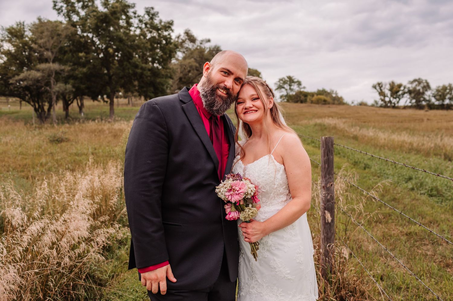 Bride and groom smiling together in a rustic field, bride holding pink bouquet, groom in navy suit with red tie.