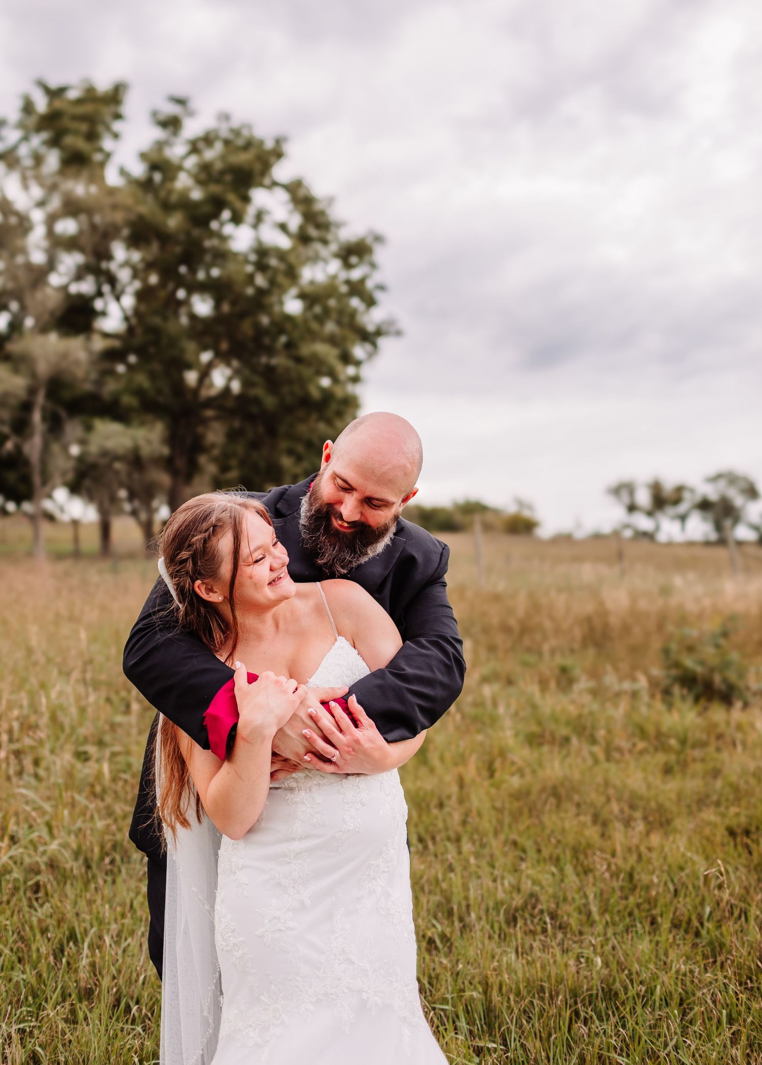 A bride and groom embrace joyfully in a grassy field, with the groom hugging her from behind on their wedding day.