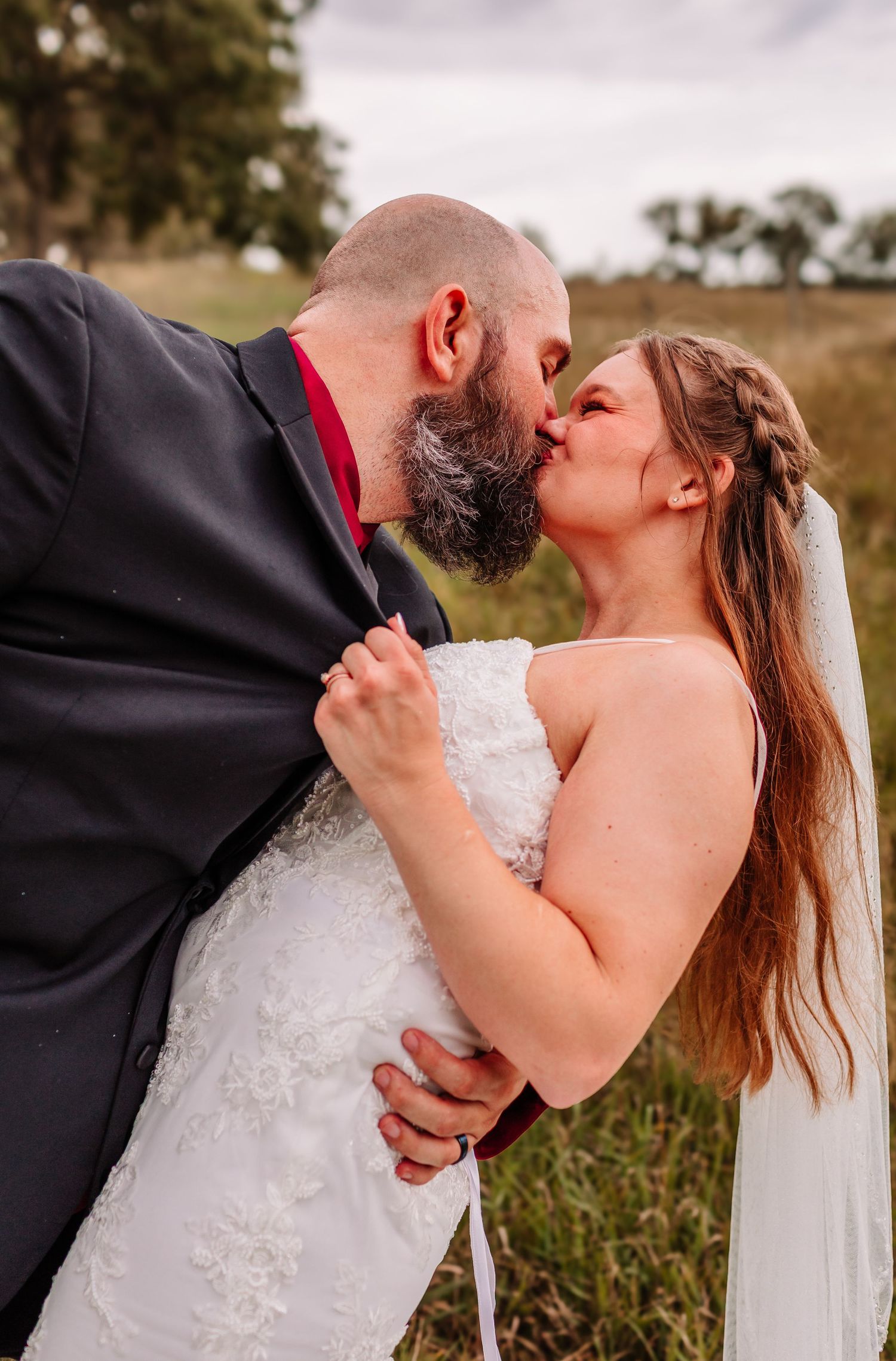 Bride and groom sharing a romantic kiss outdoors in a field, bride wearing white lace gown and veil.