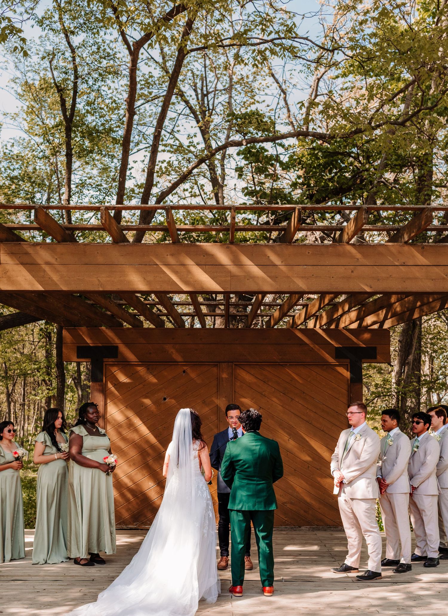 Outdoor wedding ceremony under a wooden pergola with bridal party in sage green and groom in emerald suit.