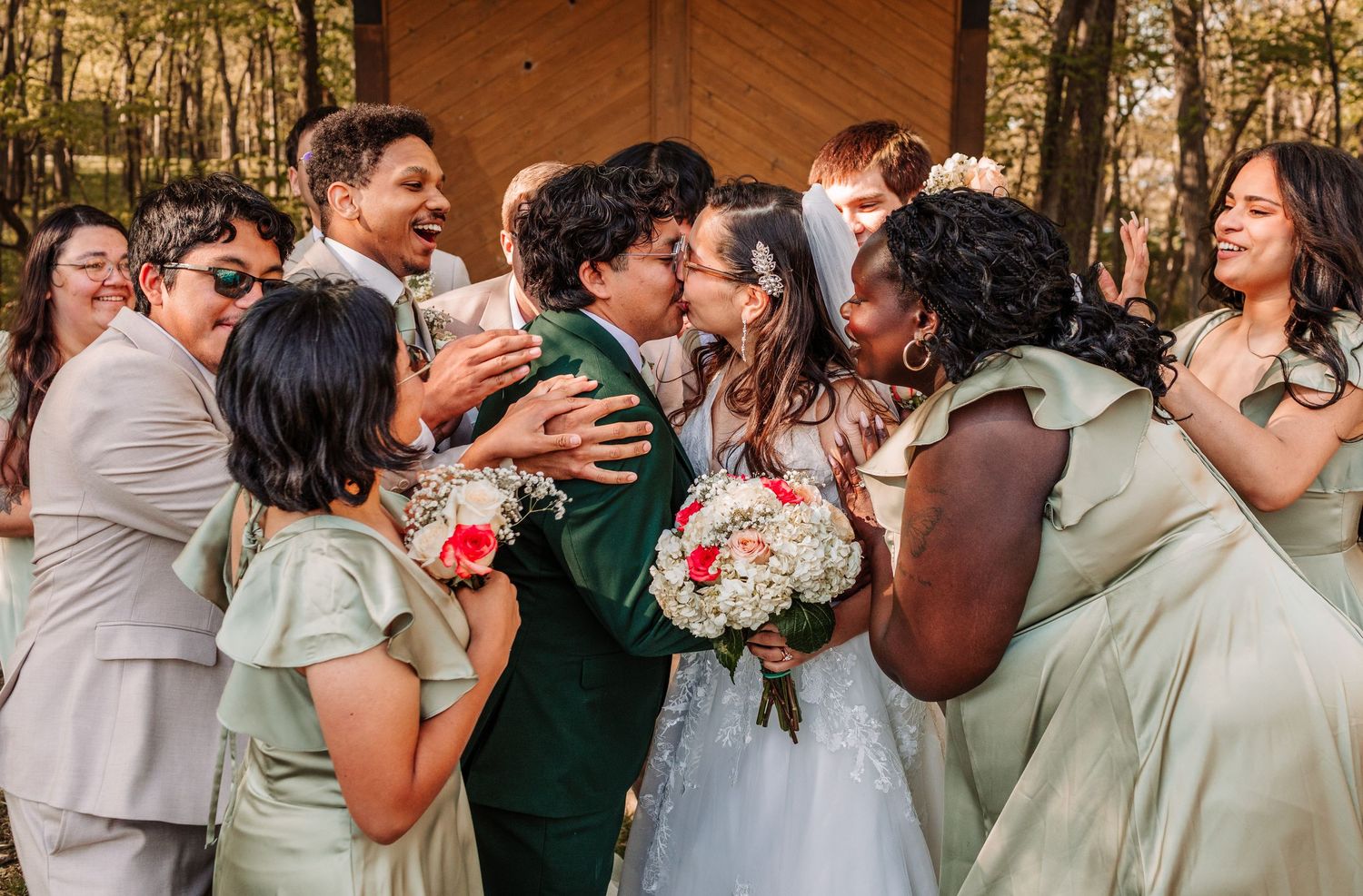 Wedding party celebrates as couple shares a kiss outdoors, surrounded by bridesmaids in sage green dresses.