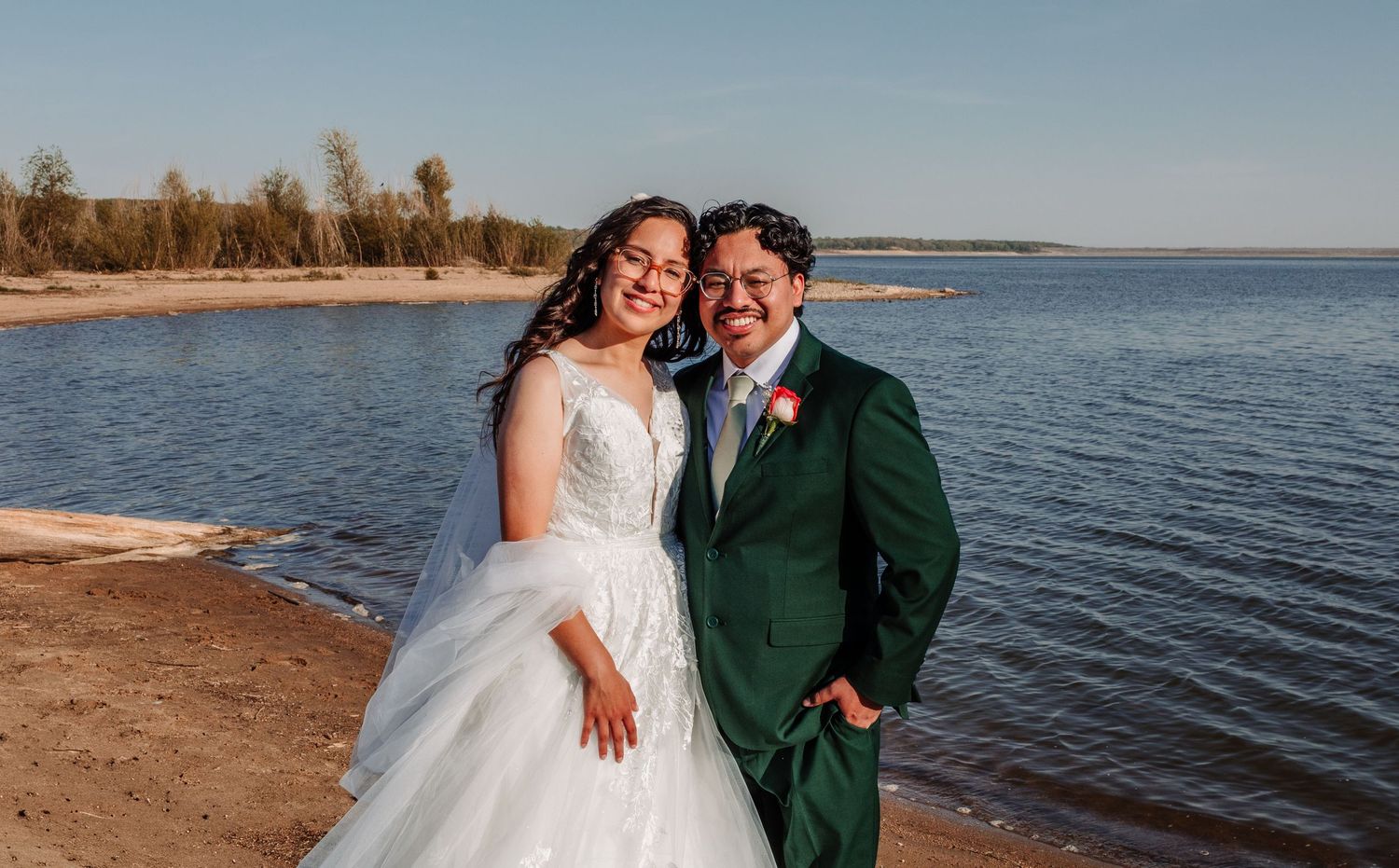 Couple in wedding attire smiling on a sandy beach by a lake, bride in white gown, groom in green suit.