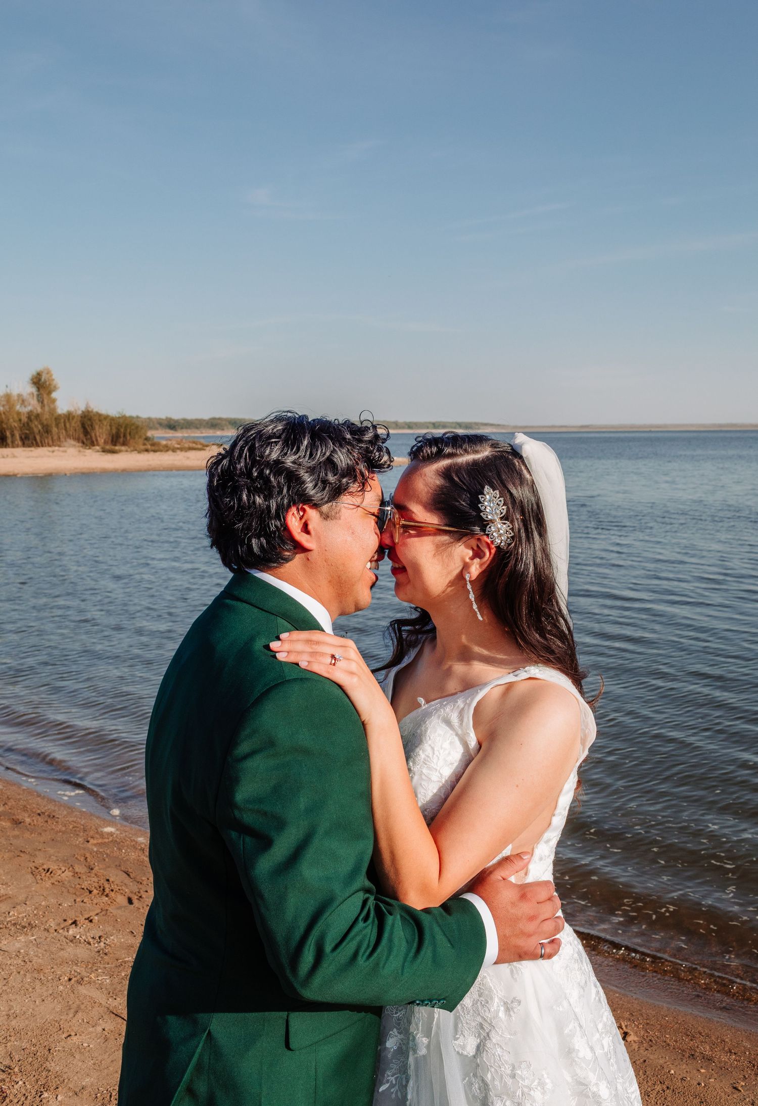 A bride and groom share a romantic moment by a lakeside, dressed in a white gown and green suit on their wedding day.